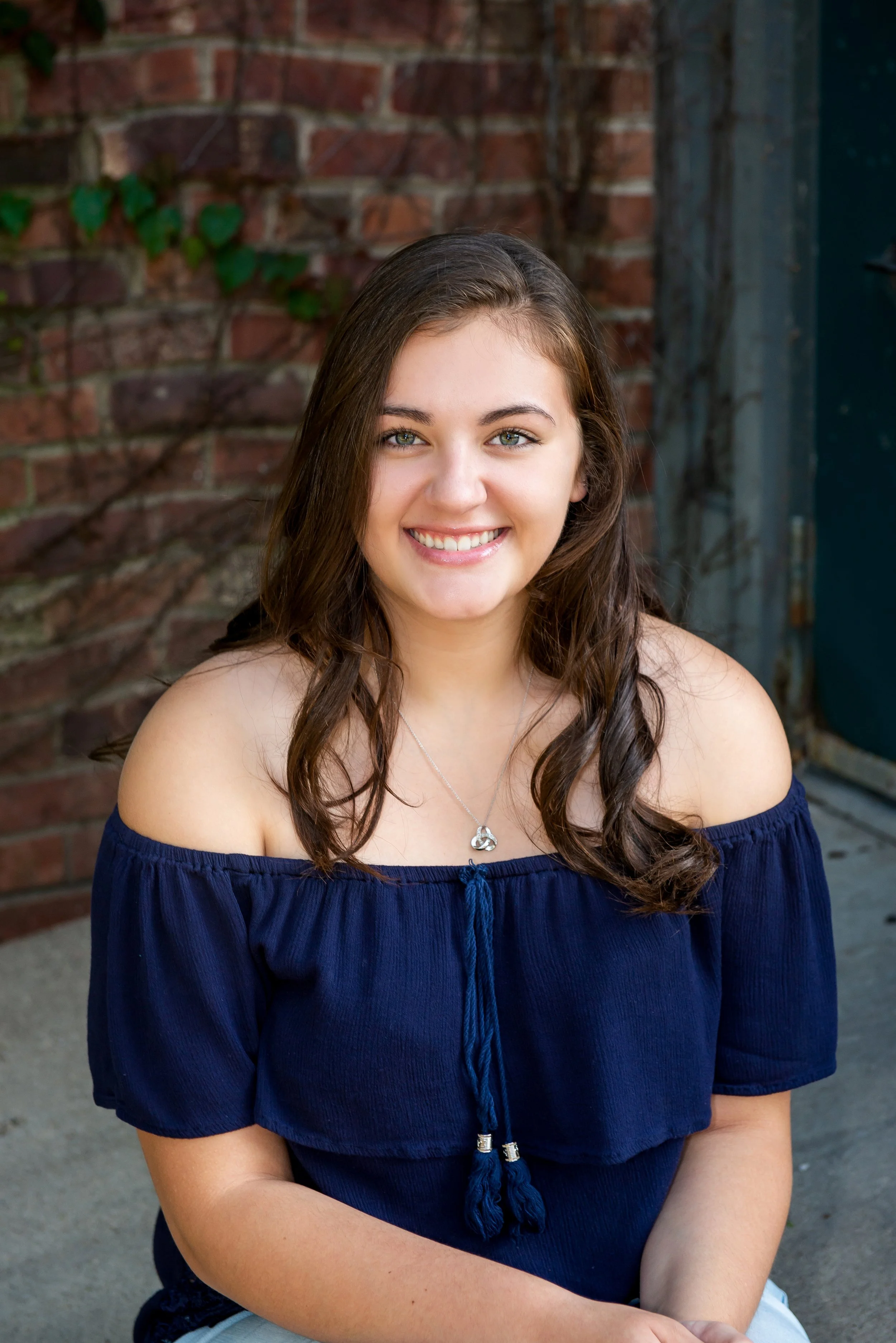 A young woman with long brown hair and green eyes smiling, wearing an off-shoulder blue top and a silver necklace, sitting outdoors against a brick wall.