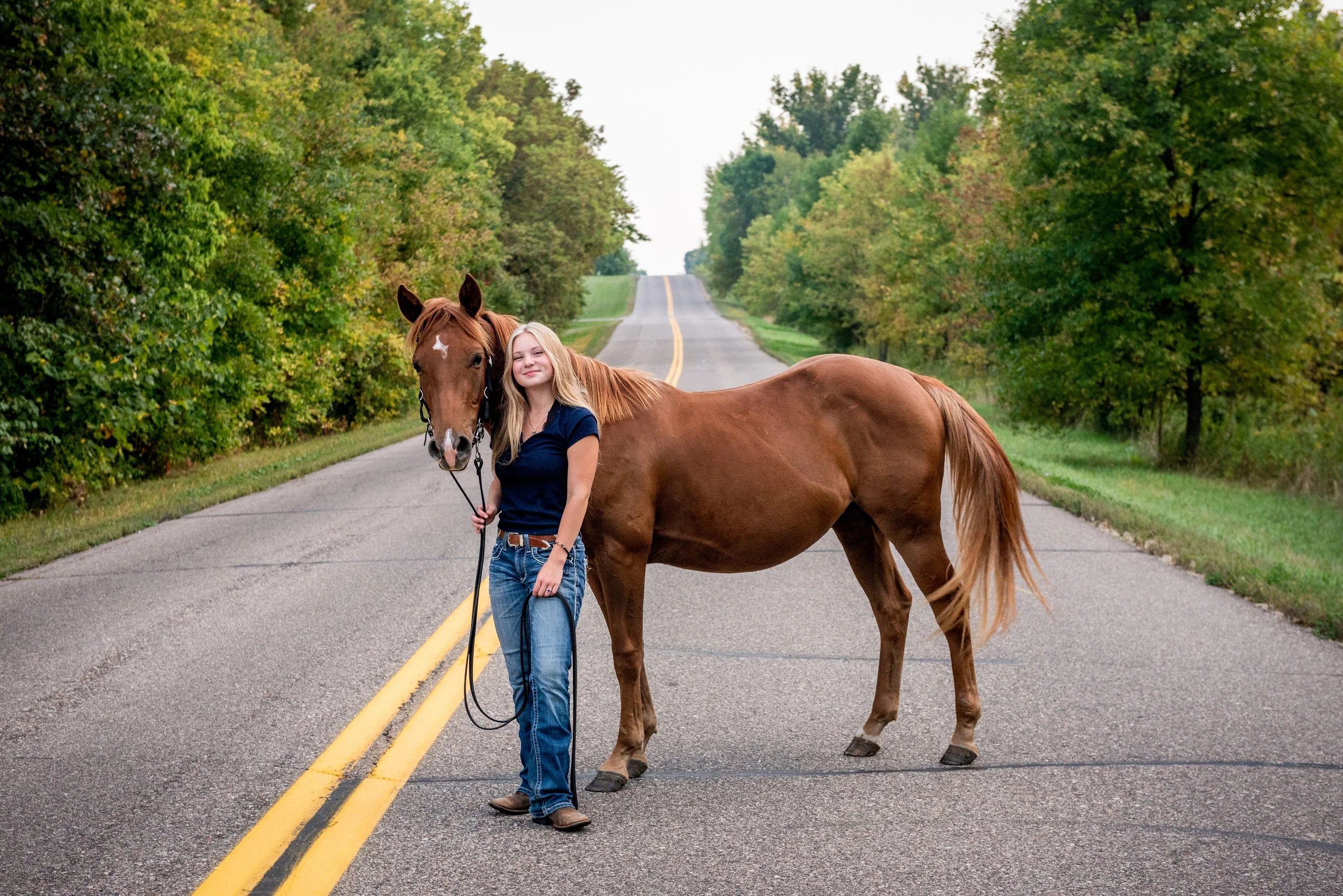 Young woman standing on a rural road holding a horse's lead, surrounded by green trees.