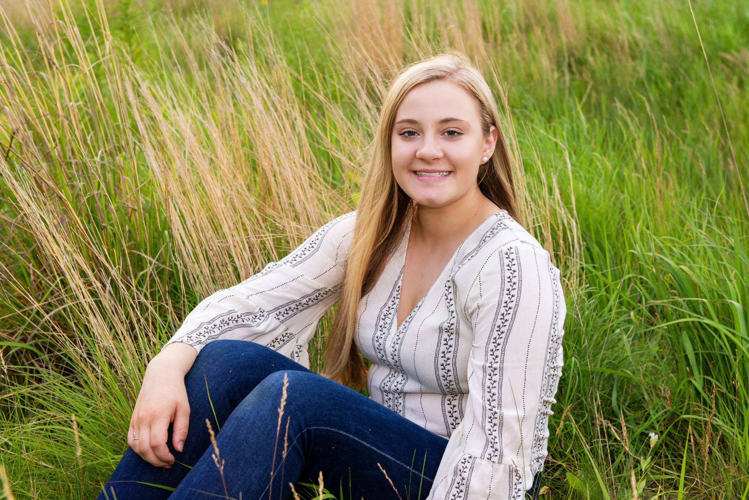 A young woman with long blonde hair, wearing a white embroidered blouse and blue jeans, sitting in a green grassy field and smiling at the camera.