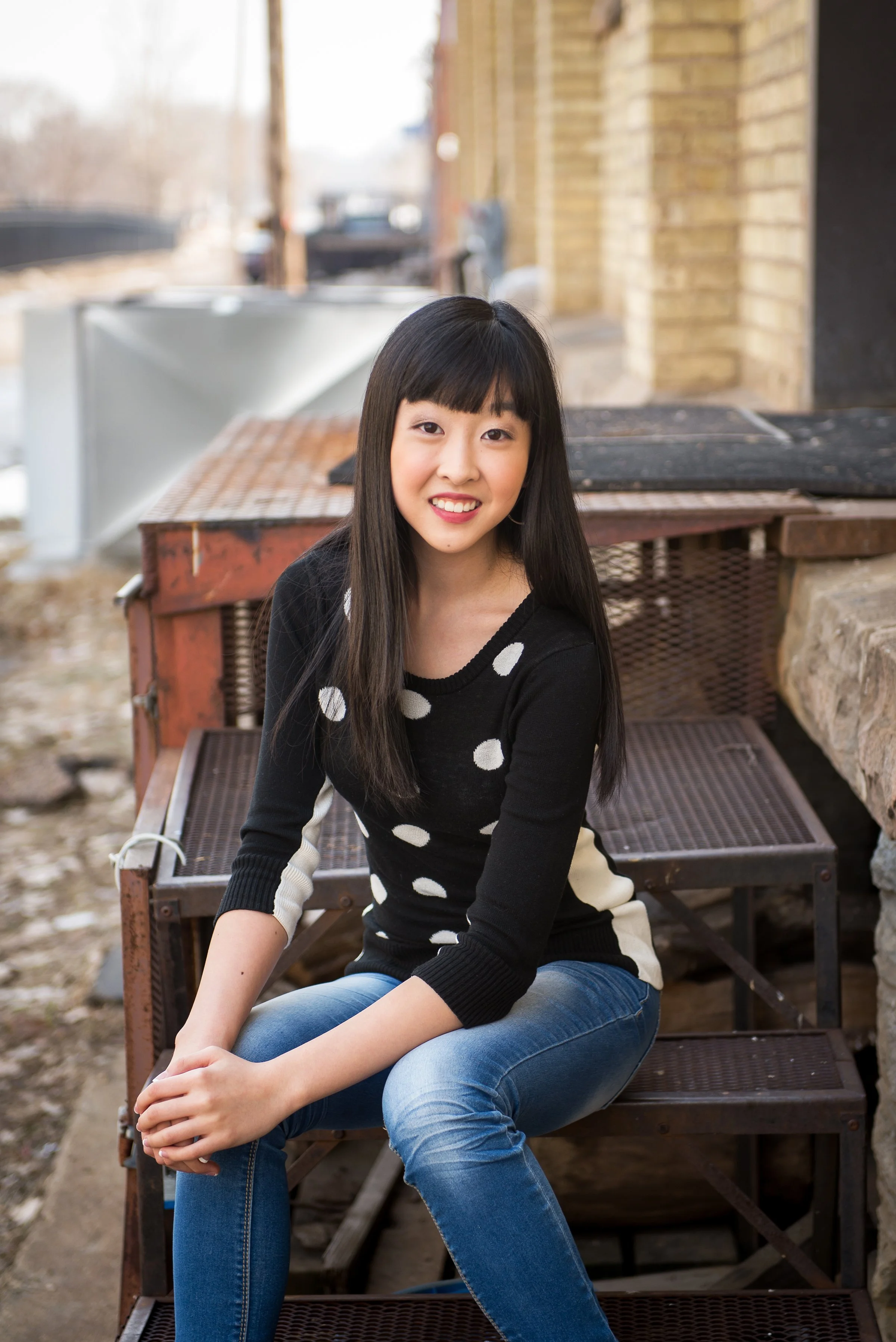 A young woman with long dark hair and a black sweater with white polka dots, sitting on a metal bench outside in front of a brick building, smiling at the camera.