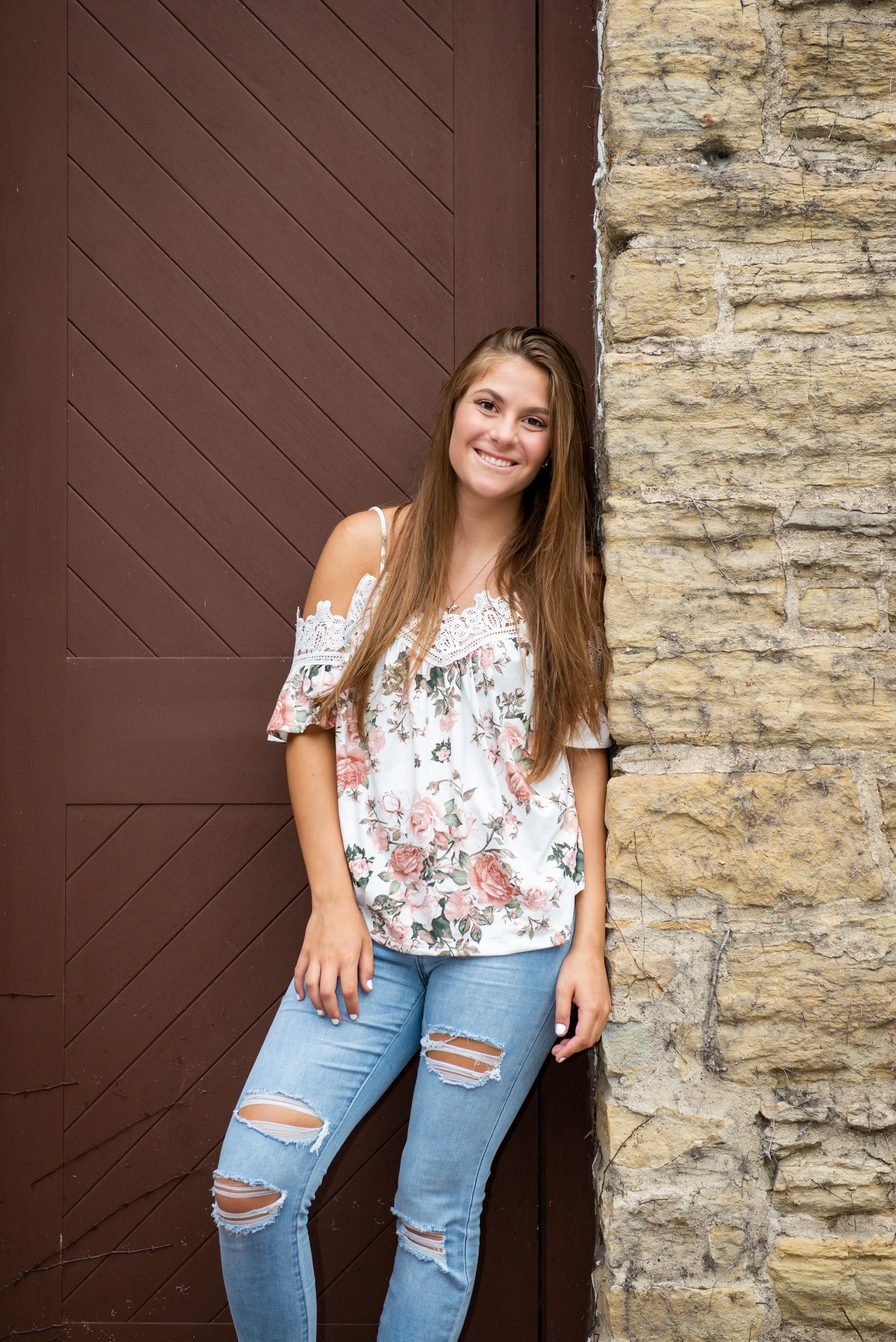 A young woman with long brown hair is smiling, standing outdoors against a stone wall and a wooden door. She is wearing a white, floral off-the-shoulder top and ripped blue jeans.
