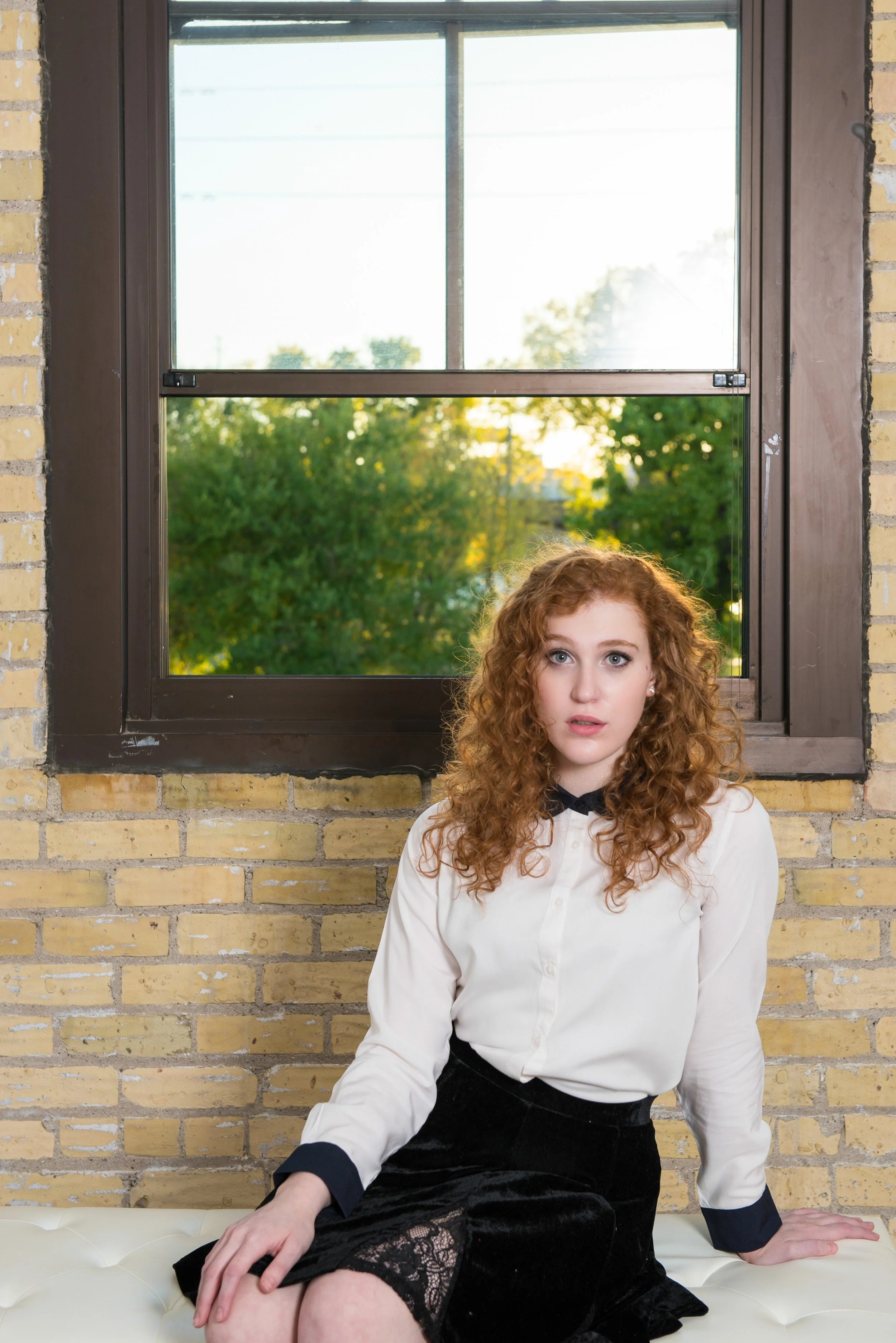 A young woman with curly red hair sitting on a white cushioned surface in front of a window with a brick wall. Outside the window, green trees are visible with sunlight shining through.