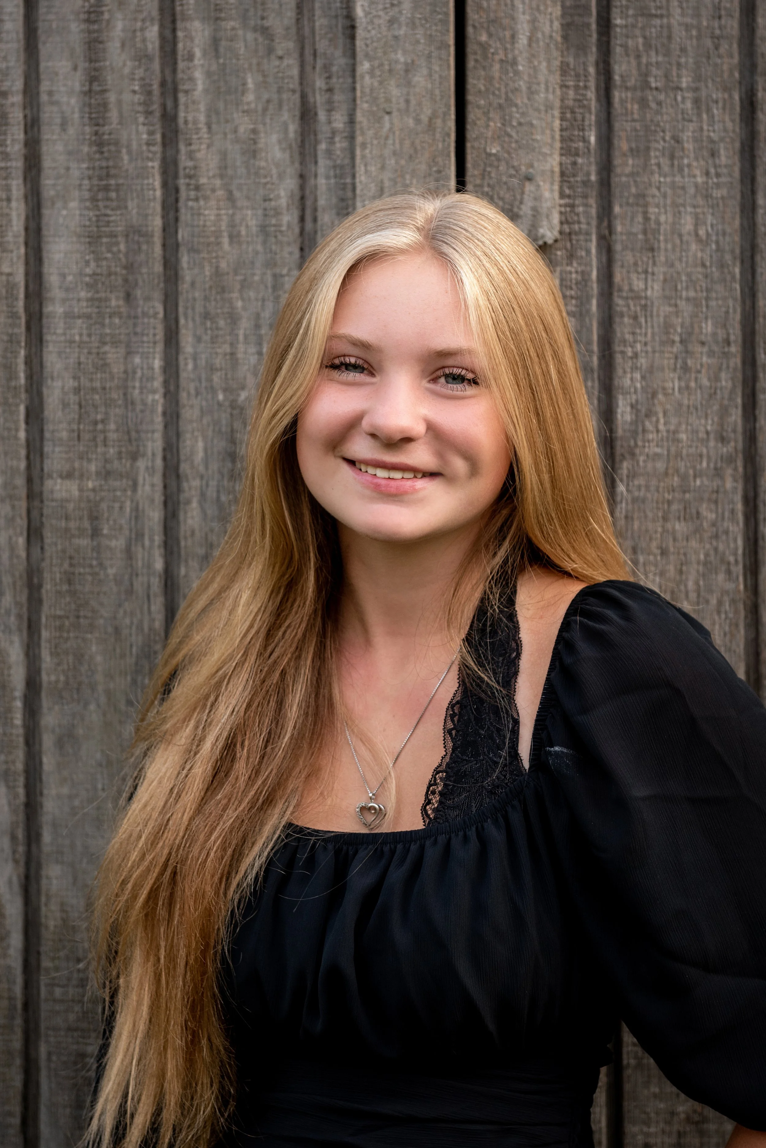 A young woman with long blonde hair smiling in front of a wooden fence.