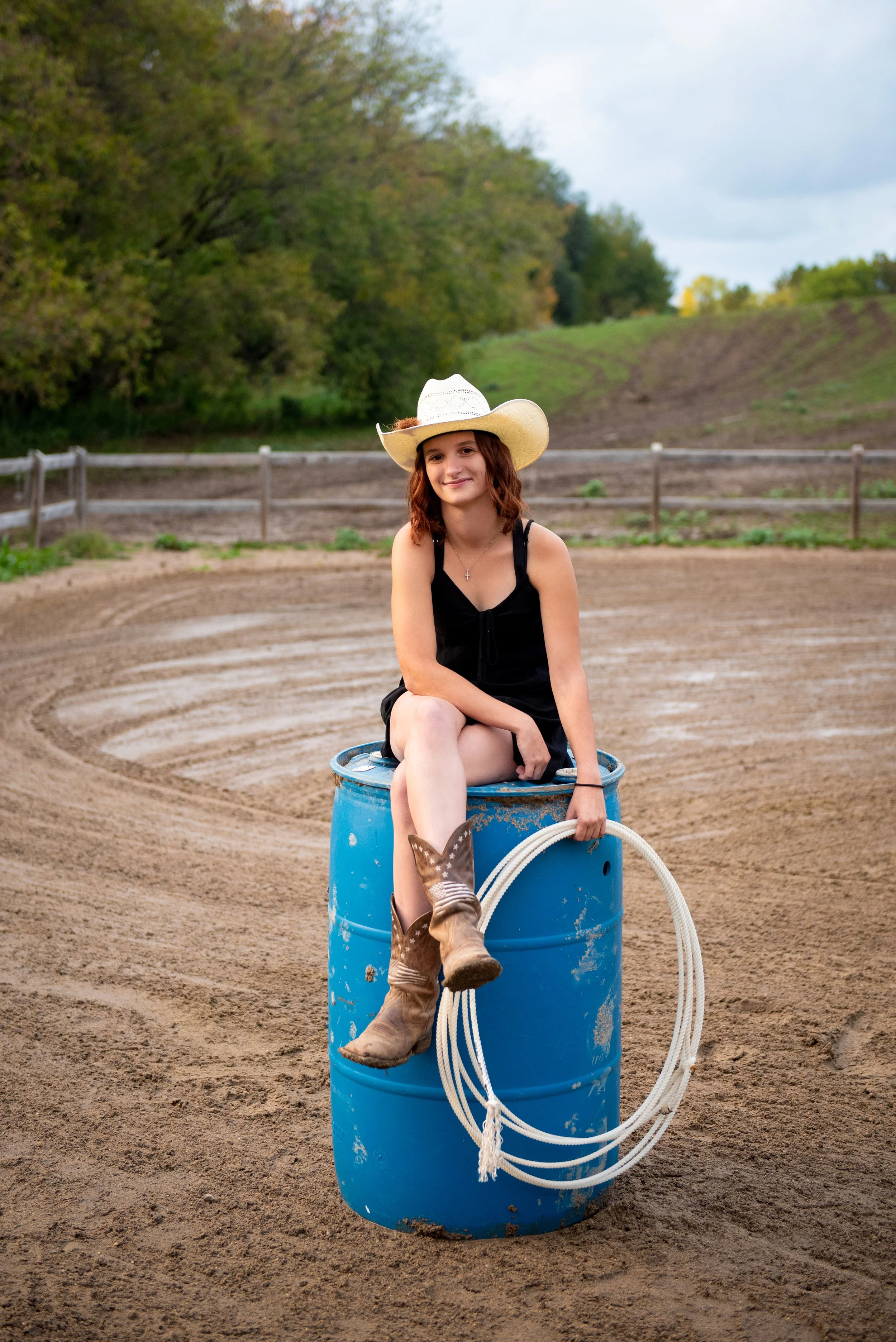 A teenage girl with red hair, wearing a cowboy hat, black dress, and cowboy boots, sitting on a blue barrel on a dirt rodeo or arena. She is holding a lasso and smiling, with a background of fencing and green trees.