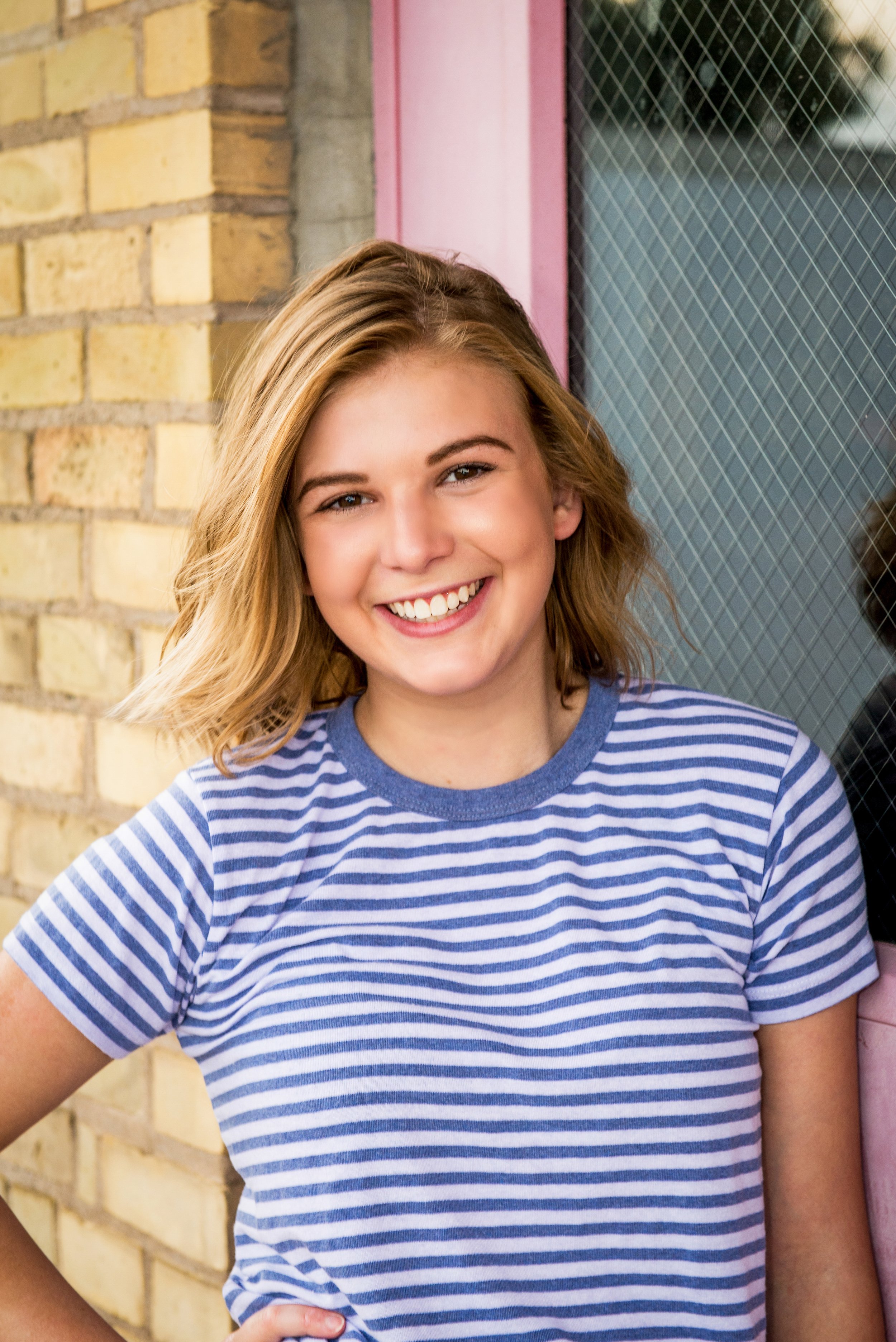 A young woman with blonde hair, smiling, wearing a blue and white striped T-shirt, standing outdoors against a brick wall and a window.