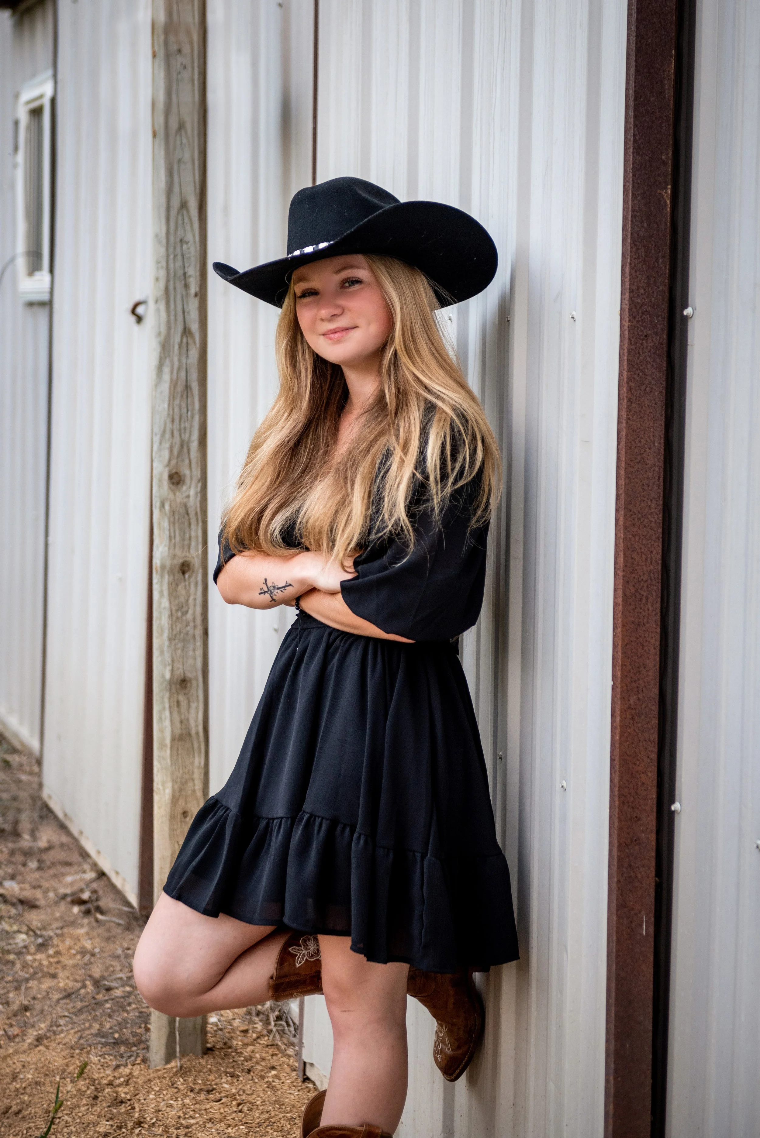 Young girl with long blonde hair wearing a black cowboy hat, black dress, and brown cowboy boots, standing with arms crossed against a metal siding wall outdoors.