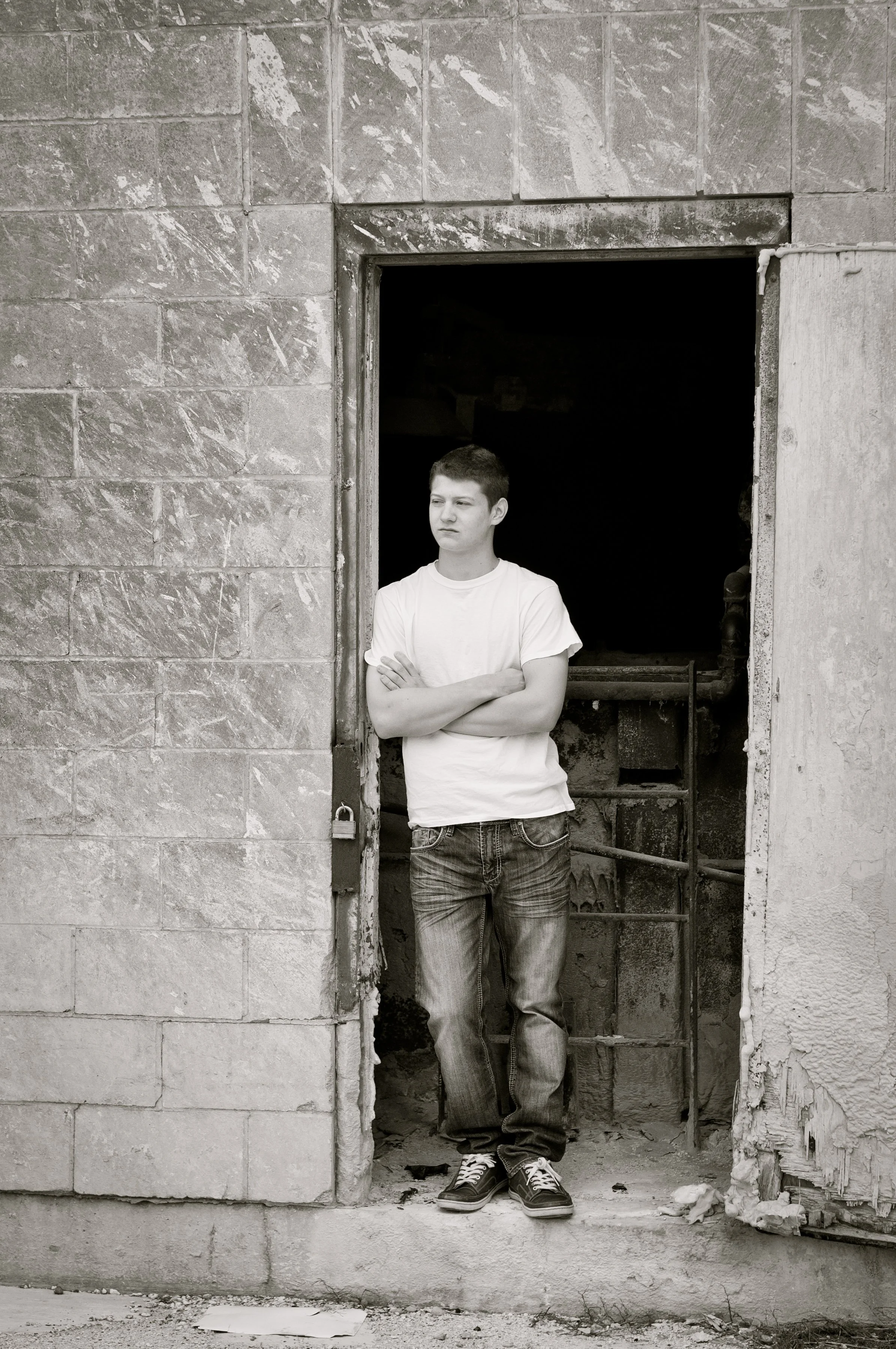 A young man with crossed arms standing in the doorway of a partially constructed building, wearing a white t-shirt, jeans, and sneakers. The building is made of concrete blocks and appears to be unfinished.