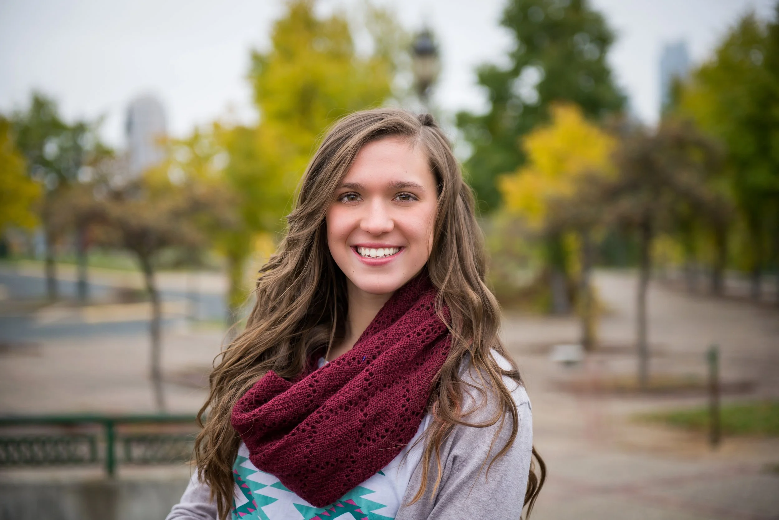 A young woman with long wavy brown hair and a bright smile, wearing a maroon knitted scarf and a light-colored top with a geometric pattern, standing outdoors in a park with trees showing fall foliage.