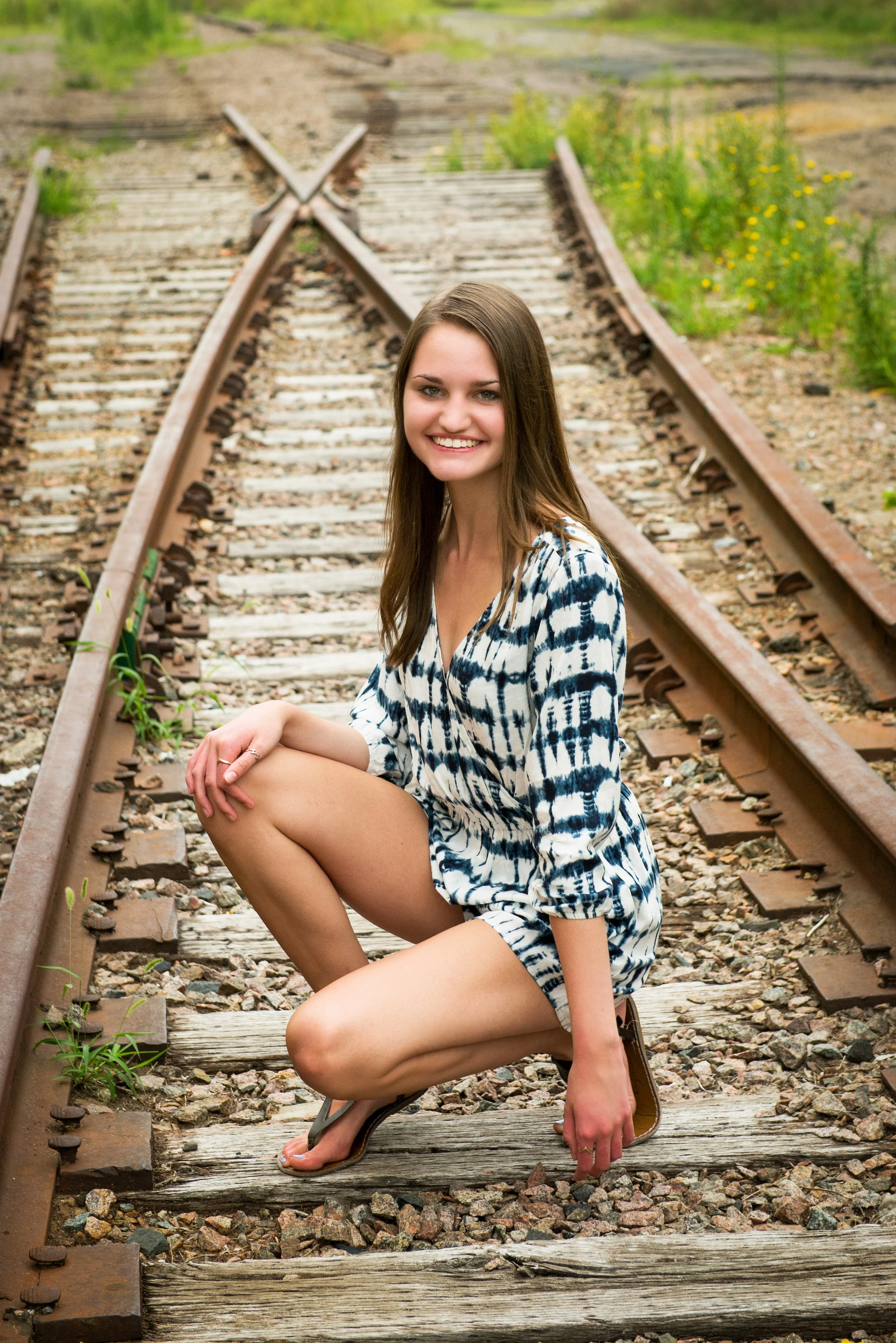 A young woman crouching on abandoned train tracks outdoors, smiling, wearing a patterned dress and flip-flops, with greenery in the background.