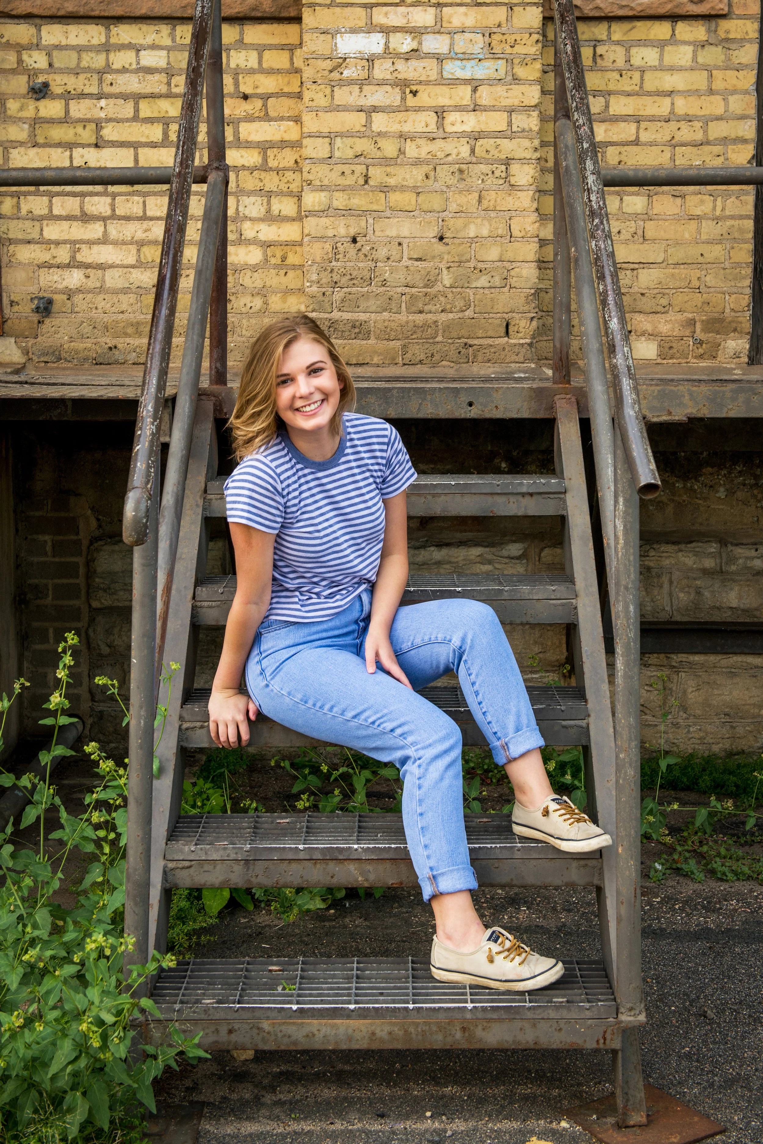A young woman with shoulder-length blonde hair, wearing a blue and white striped t-shirt, blue jeans, and beige sneakers, sitting on metal stairs outside in front of a brick wall, smiling at the camera.