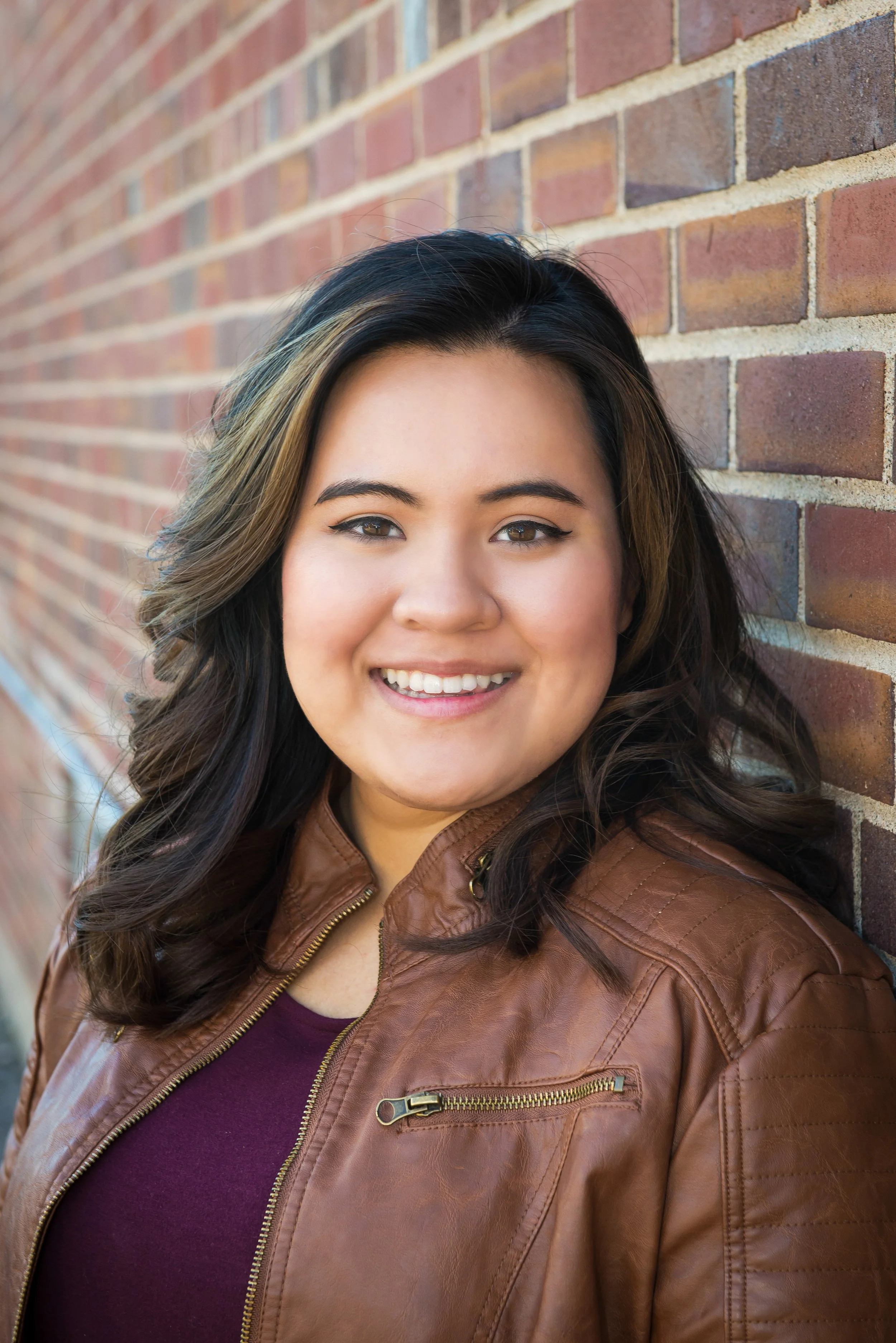 A woman with dark, wavy hair smiling and leaning against a brick wall outdoor shot.