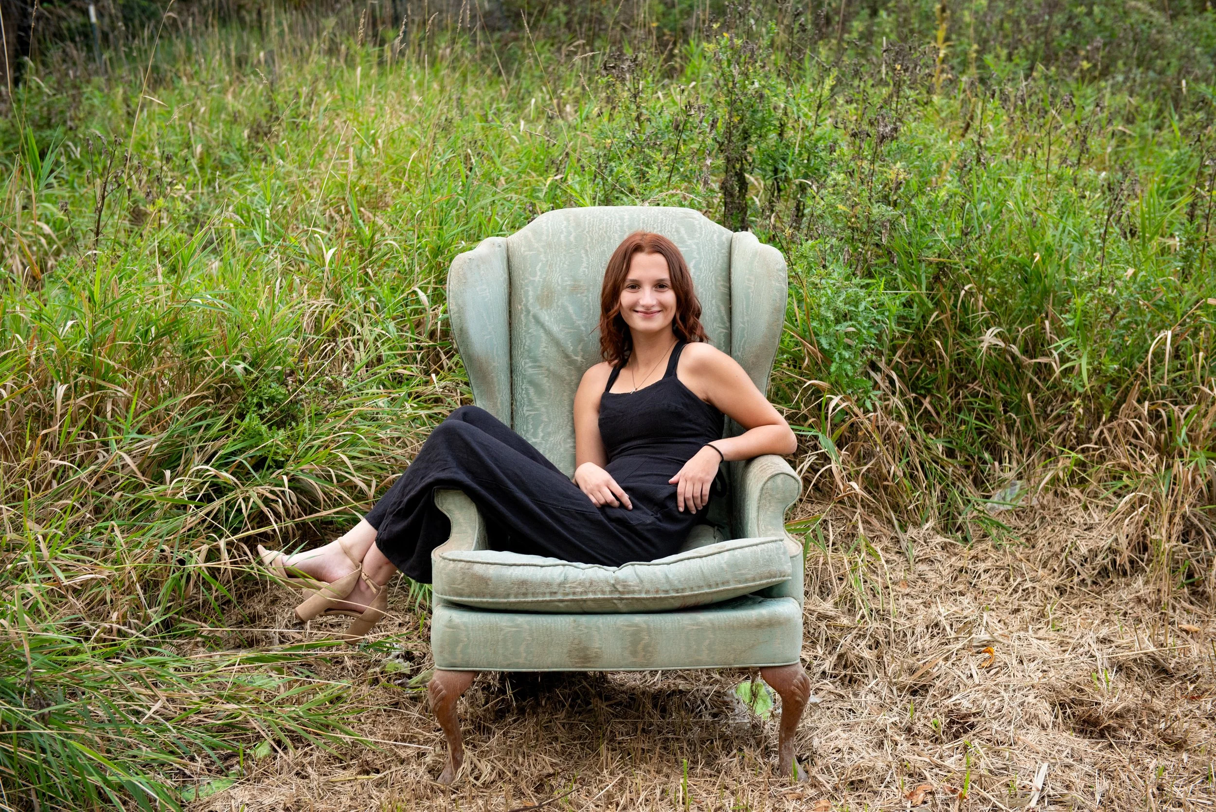 A woman with brown hair, wearing a black dress and heels, sitting on an vintage green armchair outdoors amidst tall grass and plants, smiling at the camera.
