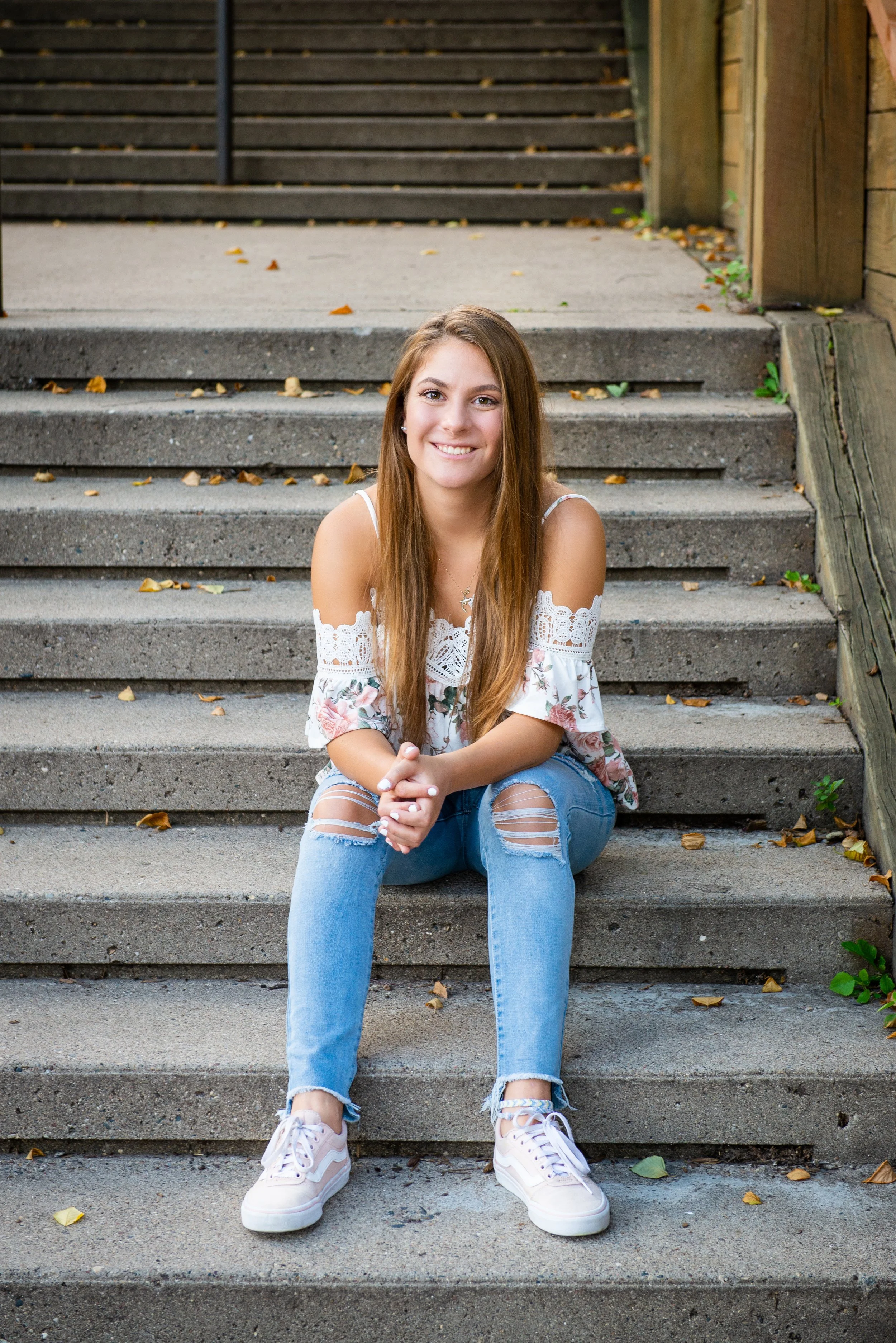 A young woman with long brown hair sits on outdoor concrete stairs, smiling at the camera, wearing a white floral off-the-shoulder top, ripped jeans, and white sneakers.