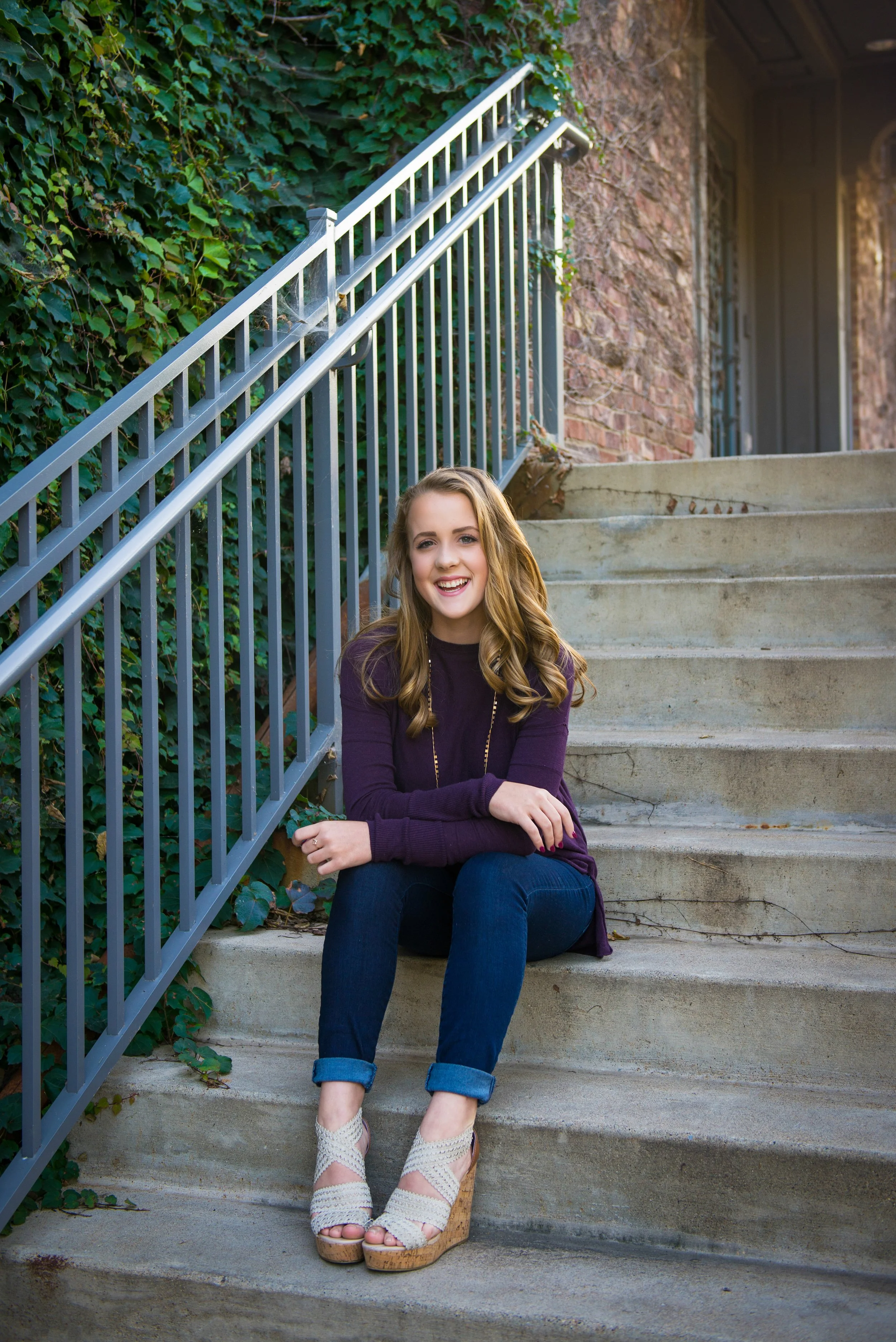 A young woman with long, wavy blonde hair sitting on outdoor concrete steps, smiling cheerfully, wearing a dark purple long-sleeve top, blue jeans, and white platform wedge sandals, with green ivy climbing on a metal railing beside her.