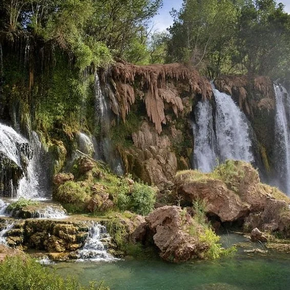 A scenic waterfall flowing over rocks and cliffs surrounded by lush green trees.