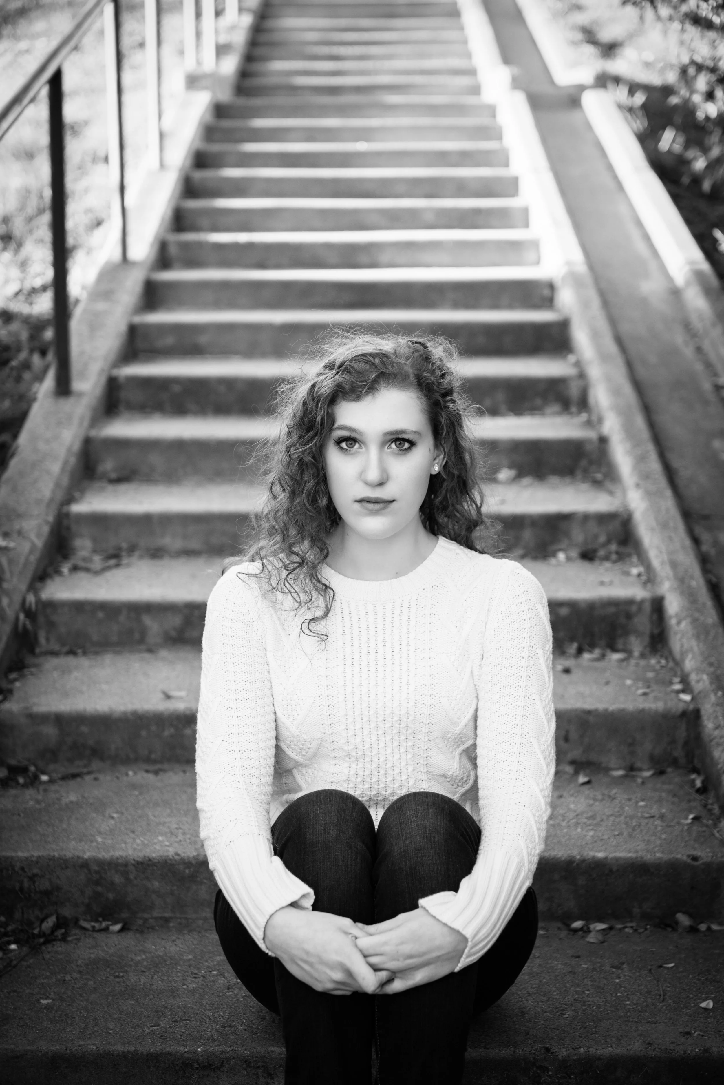 A young woman with curly hair and a white sweater sitting on the steps of an outdoor staircase.