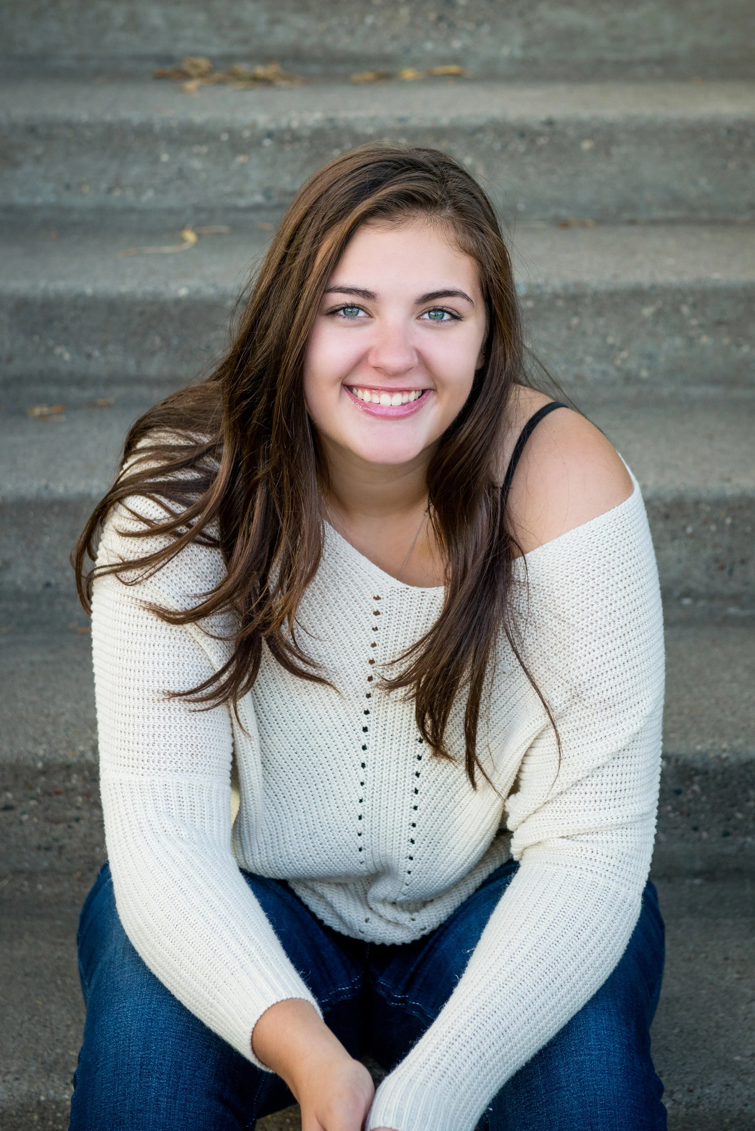 A young woman with long brown hair and blue eyes, smiling, sitting on outdoor concrete stairs, wearing a cream knitted sweater and blue jeans.