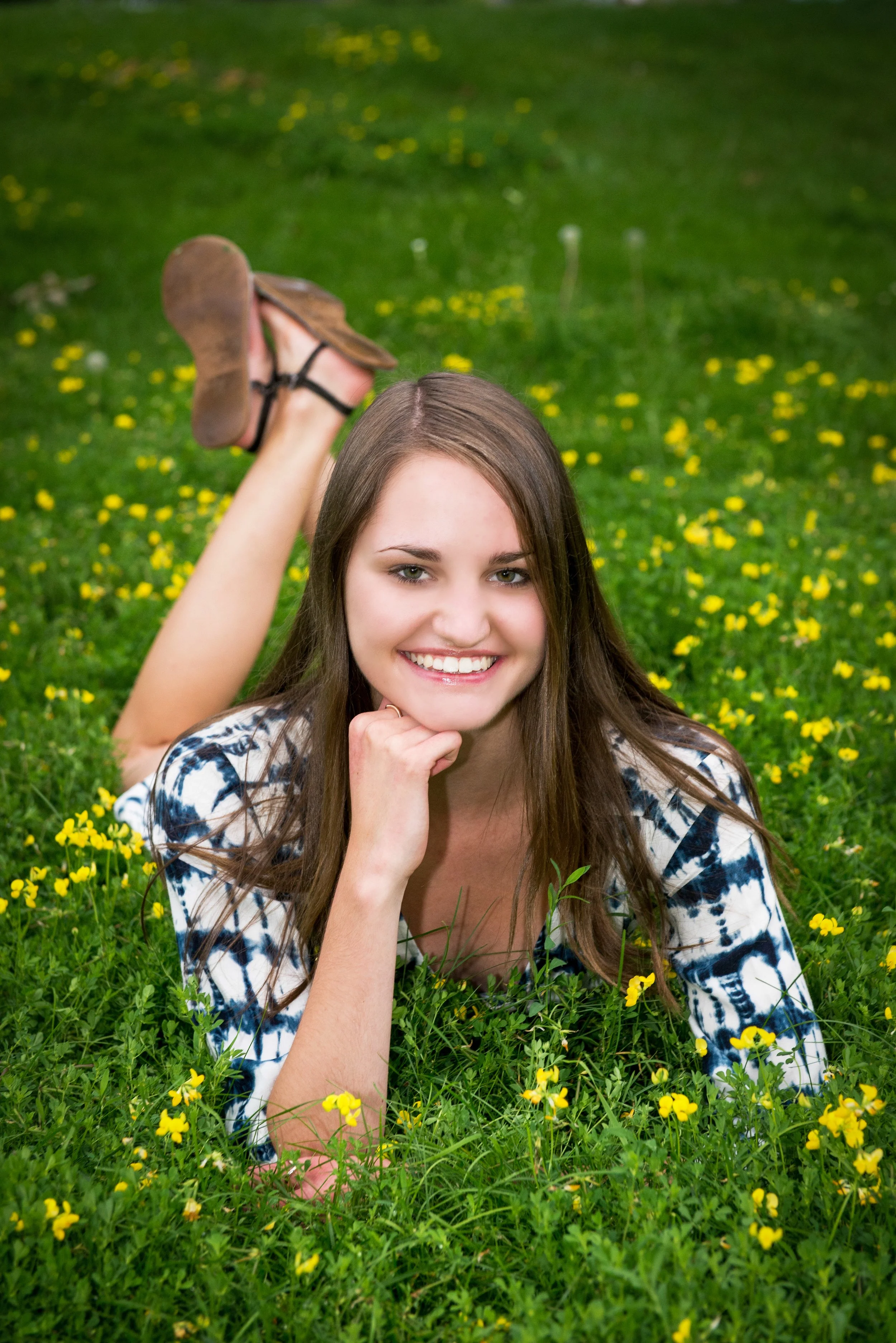 A young woman with long brown hair lies on her stomach in a field of yellow wildflowers, smiling at the camera, with her chin resting on her hand and her legs bent at the knees, feet crossed in the air.