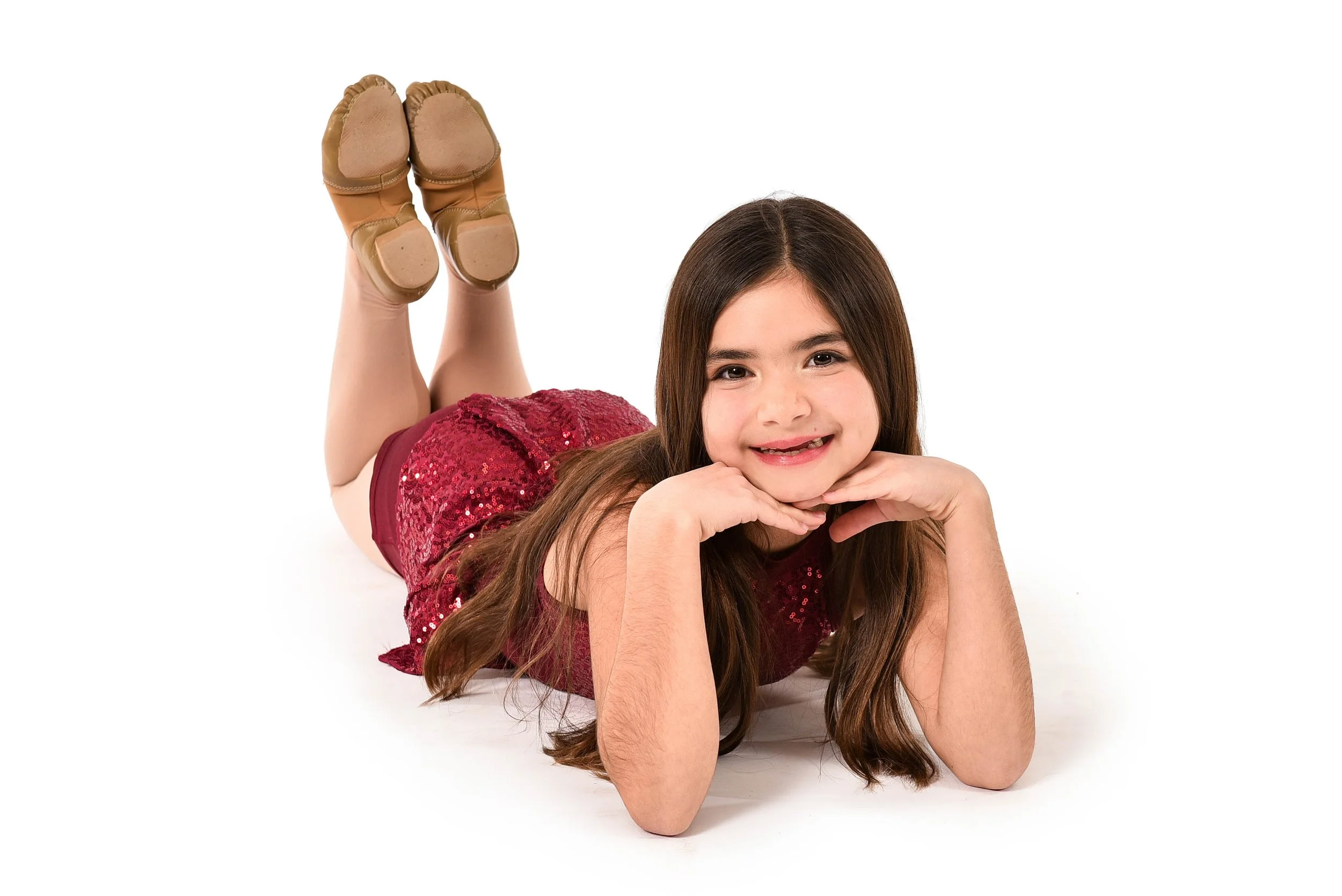 A young girl with long brown hair lying on her stomach on a white background, smiling, with her chin resting on her hands, wearing a red, sequined dress, and tan shoes.