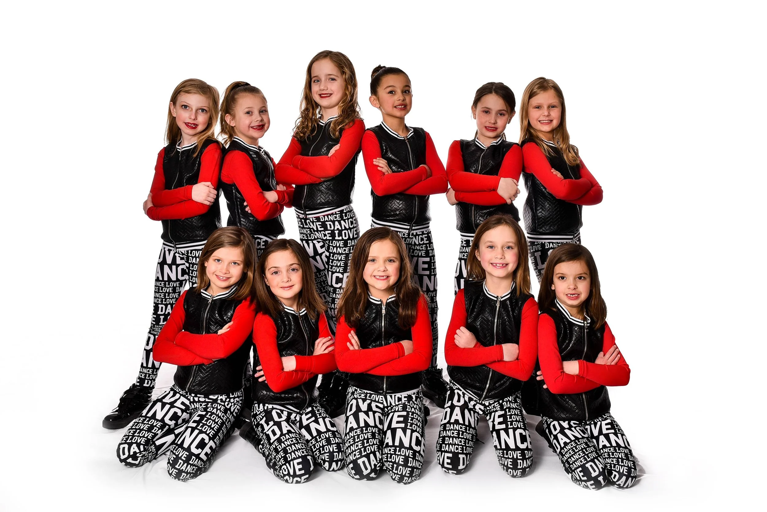 Group of ten young girls dressed in dance costumes with red and black outfits, posing with arms crossed on a white background.