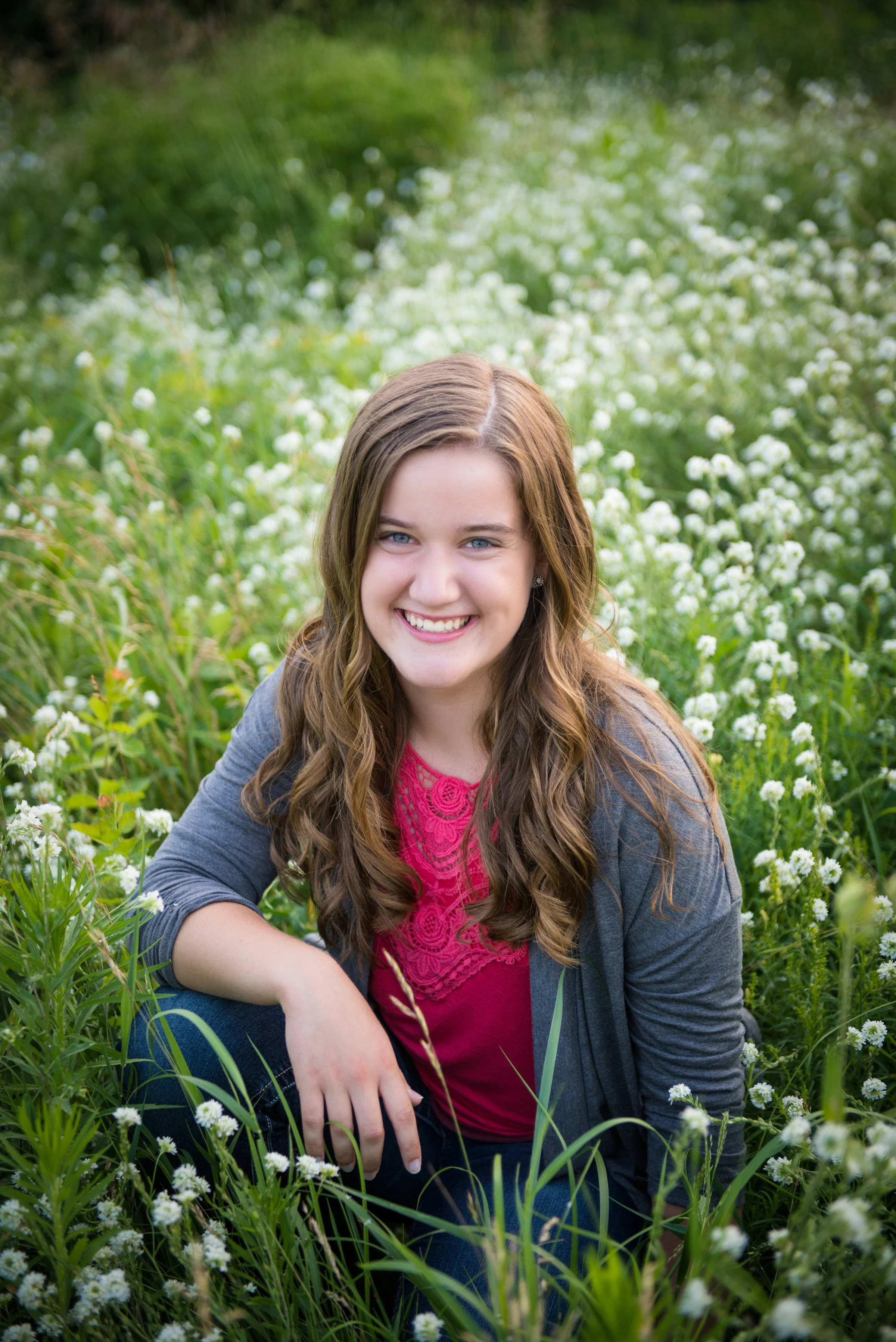 A young woman with long brown hair, wearing a pink lace top and gray jacket, sitting in a field of white flowers and green plants, smiling at the camera.