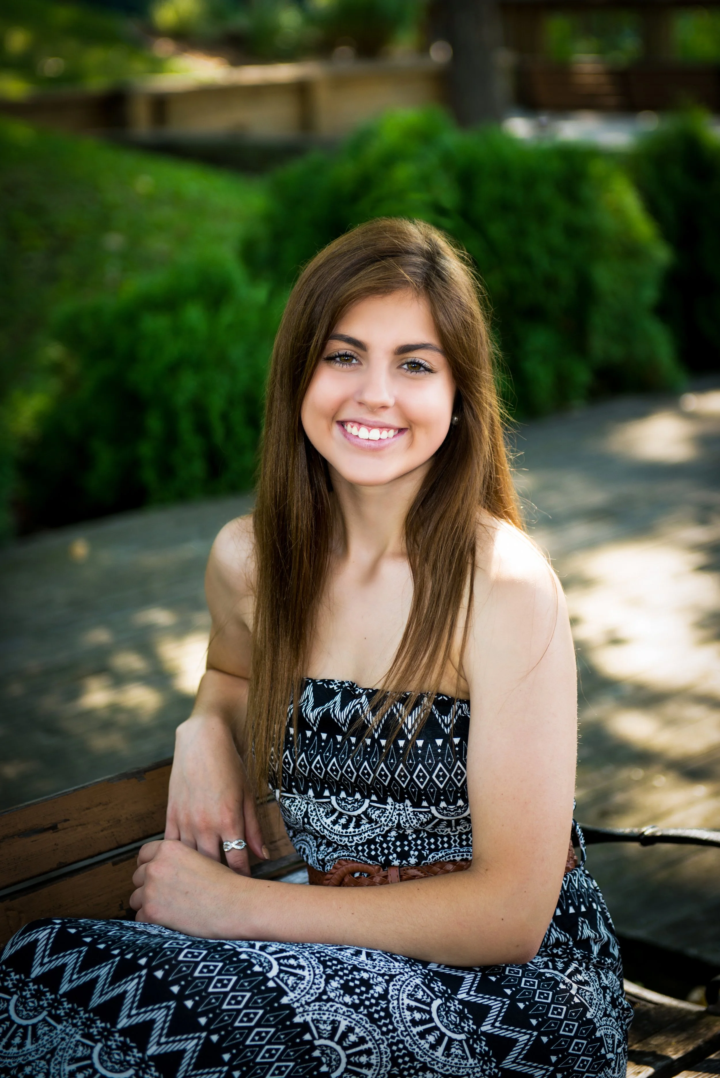 A young woman with long brown hair, wearing a black and white patterned strapless dress, sitting on a park bench outdoors, smiling.