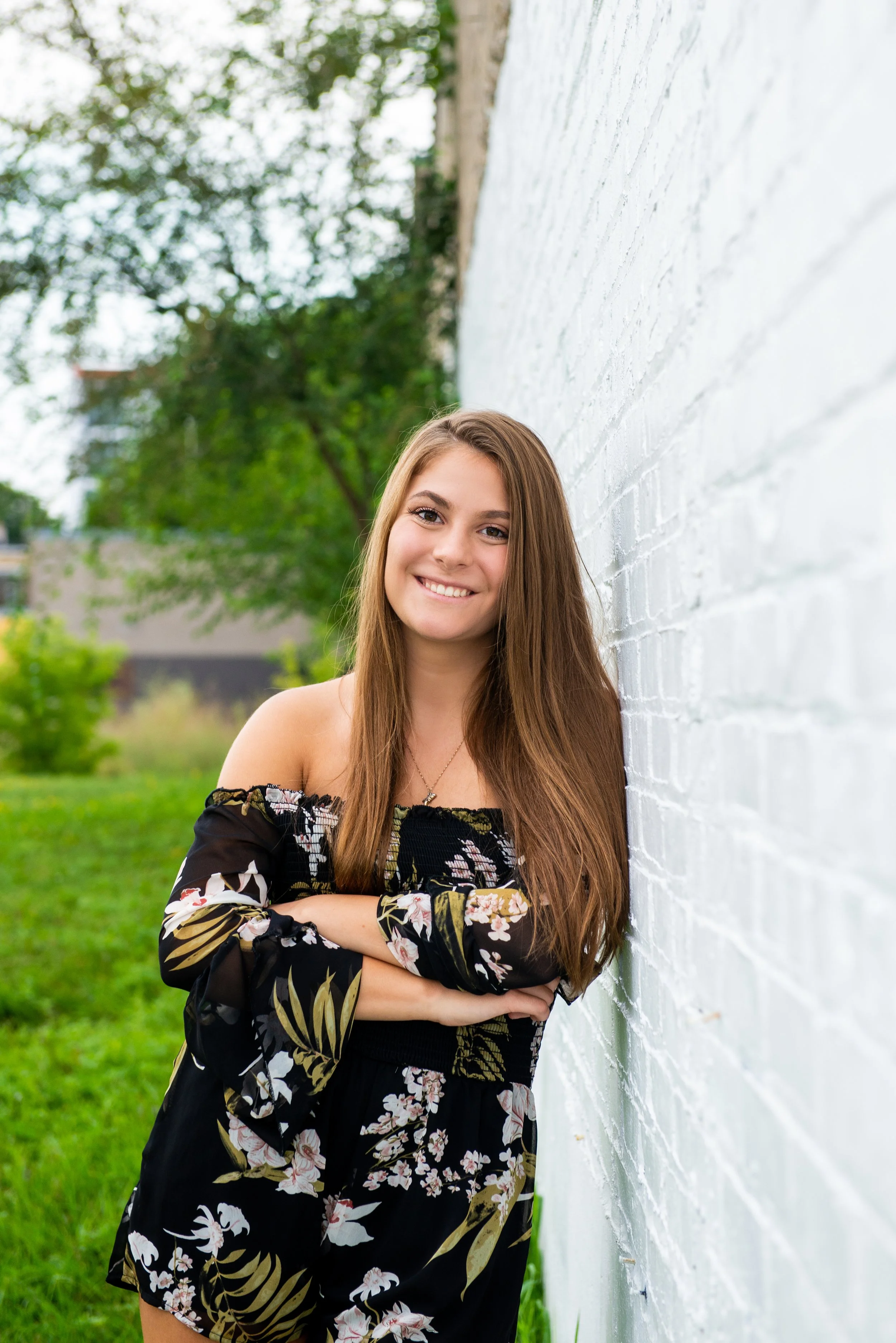 A young woman with long brown hair, smiling, wearing a black floral off-the-shoulder top and matching shorts, leaning against a white brick wall outdoors in a park-like setting with green trees and grass.
