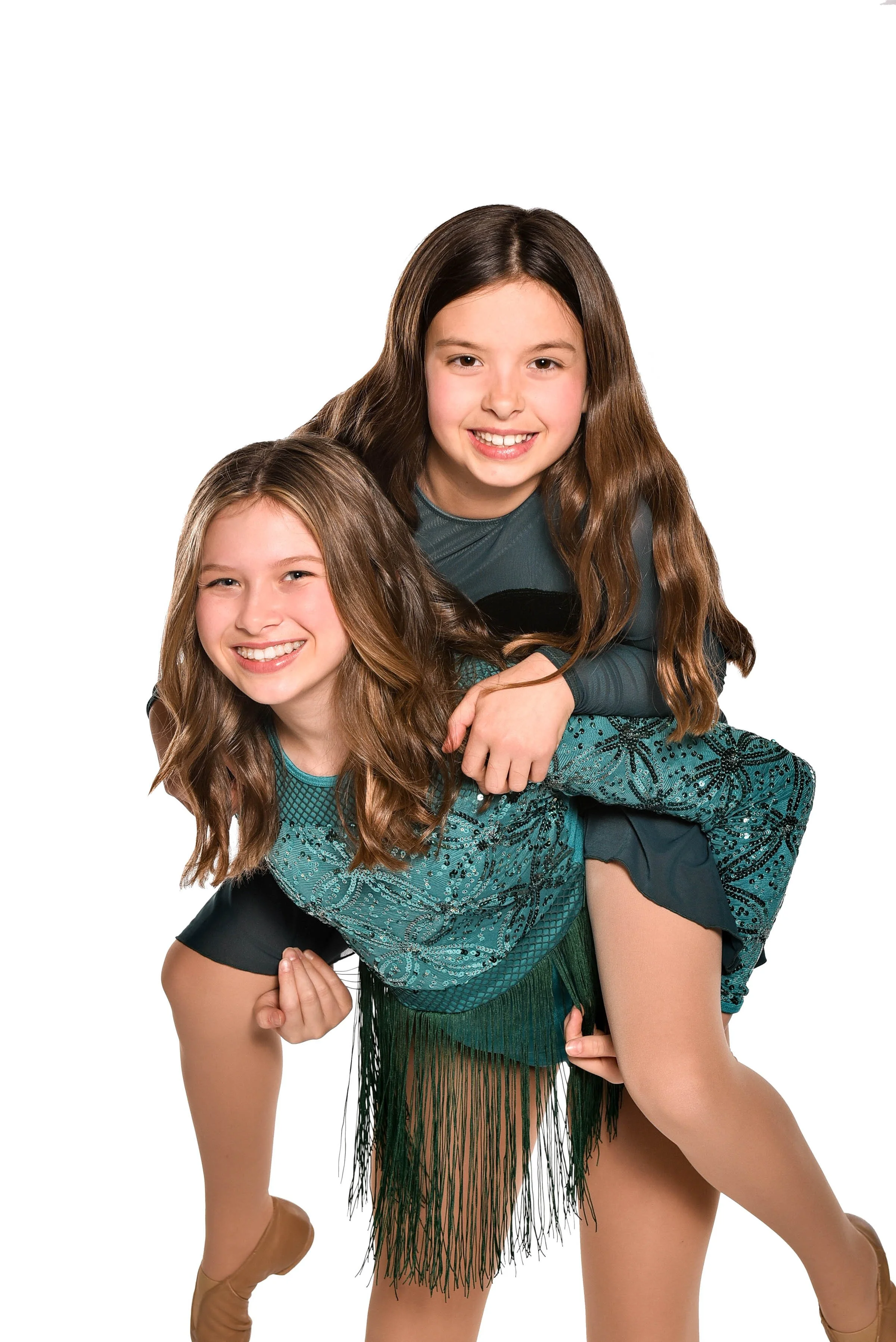 Two young girls with long brown hair, smiling, one giving a piggyback ride on the other against a white background.