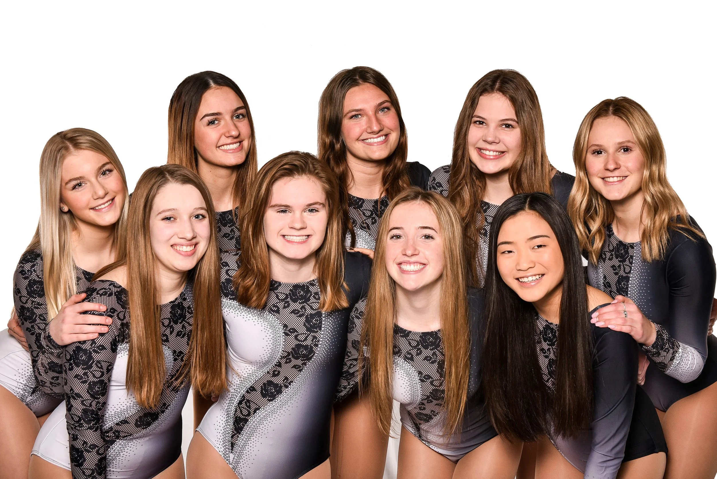 Group of ten young female gymnasts in matching black and gray leotards smiling and posing together against a white background.