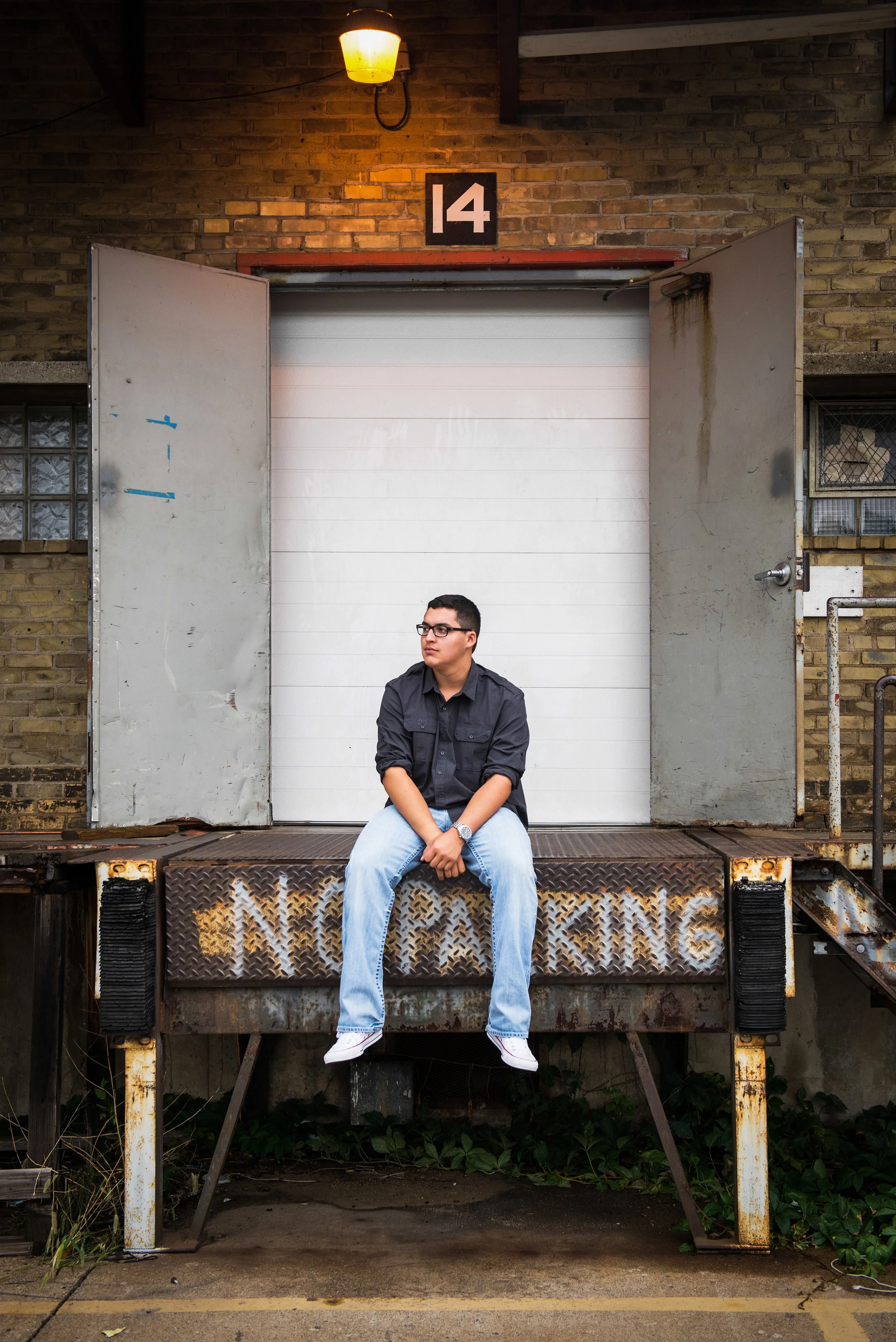 Young man sitting on a rusted loading dock with open gray doors, brick wall in the background, and a streetlamp illuminating. The dock has the words 'NO PARKING' spray-painted on it.