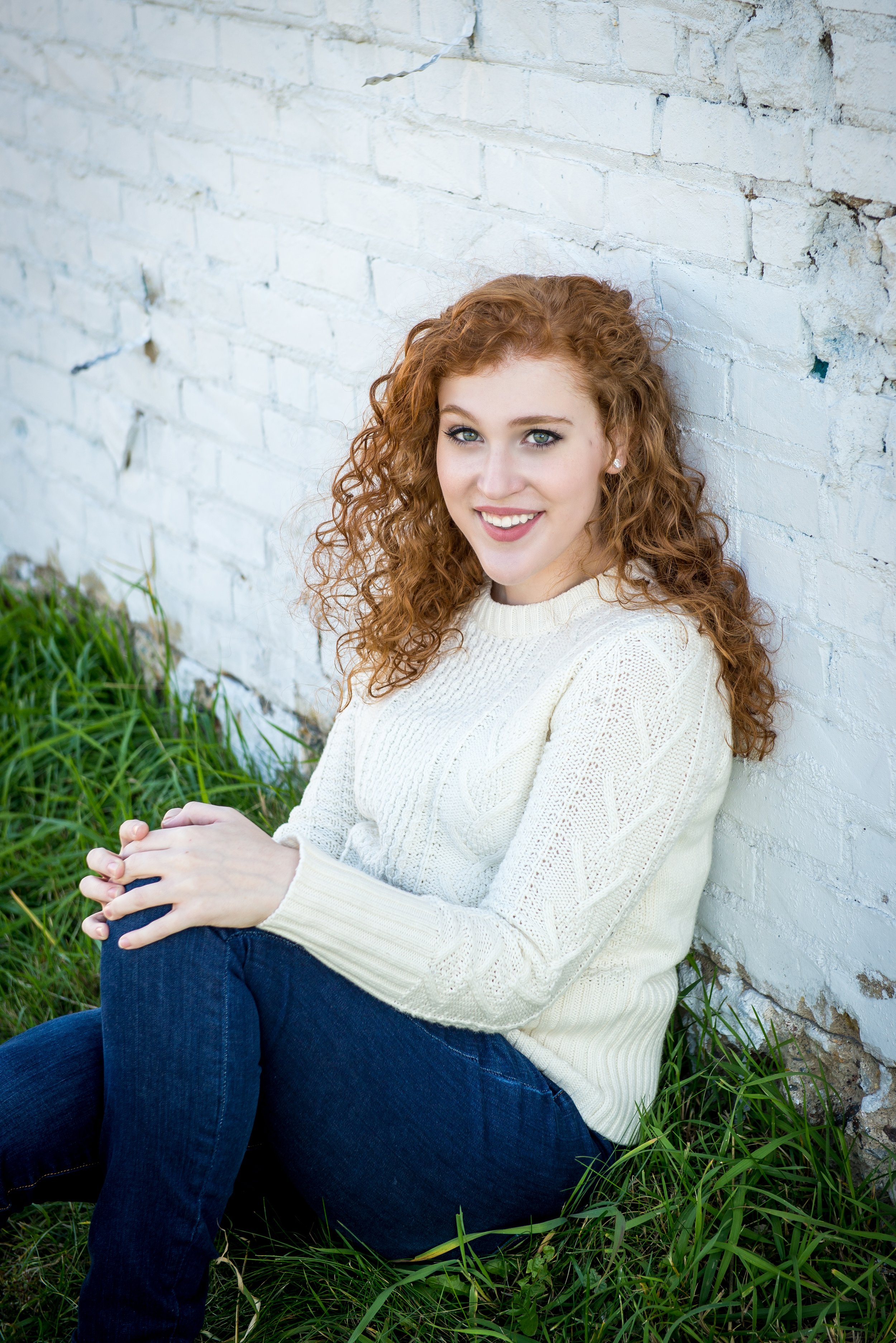 Young woman with curly red hair and blue eyes sitting outdoors against a white brick wall, smiling at the camera, wearing a cream-knit sweater and jeans, surrounded by green grass.