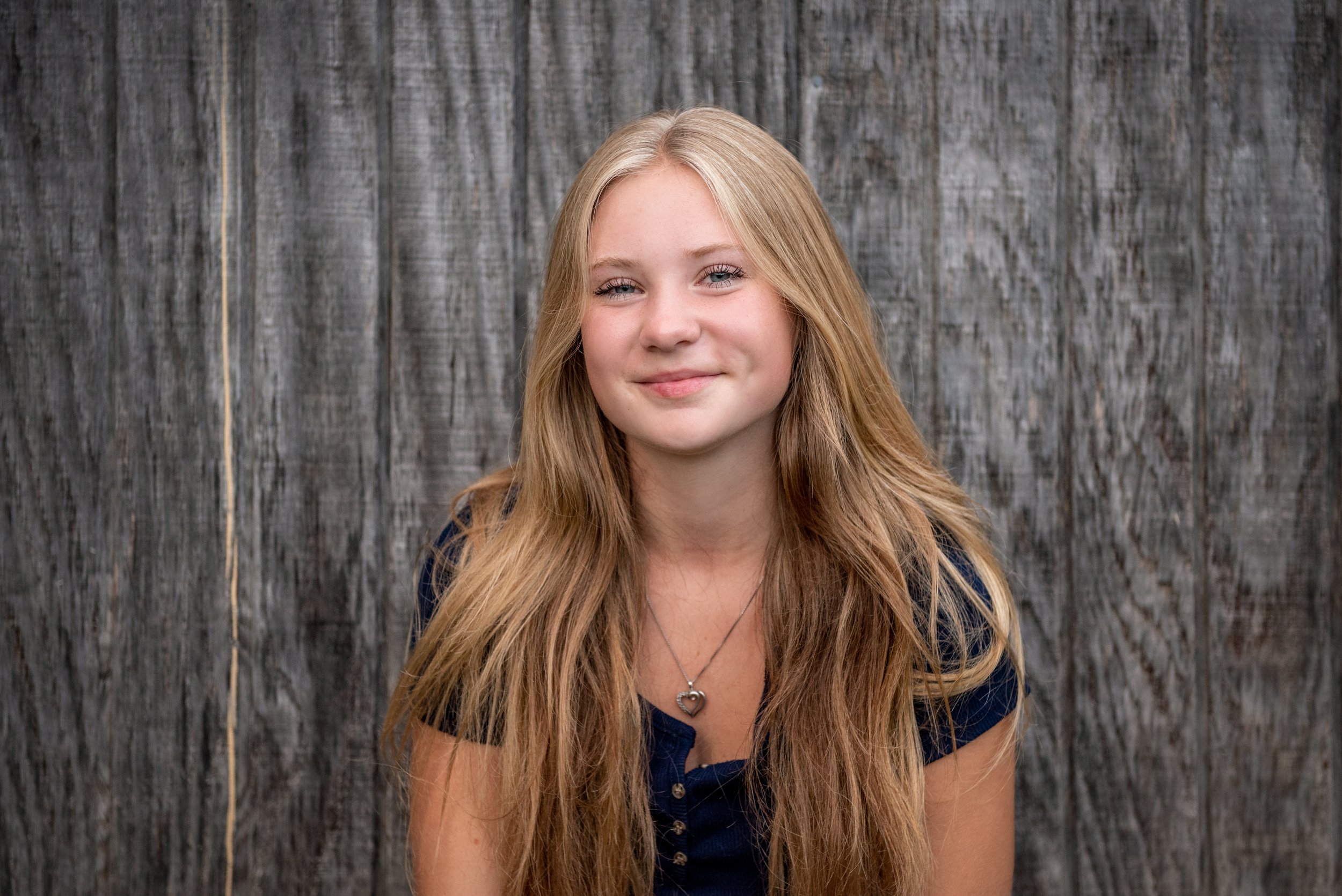 Young girl with long blonde hair, wearing a navy blue top and a heart-shaped necklace, smiling in front of a wooden fence background.