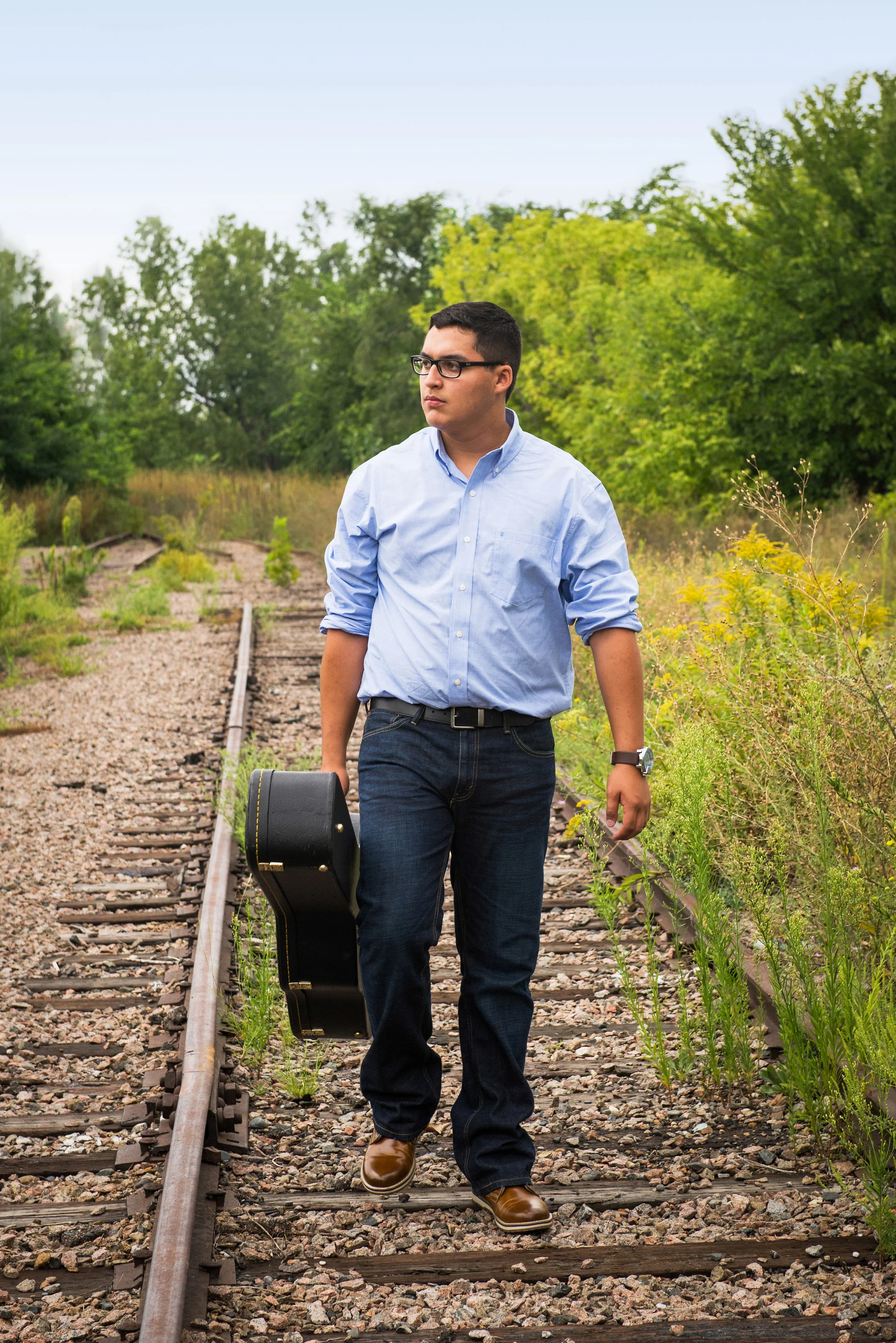 A young man wearing glasses, a light blue button-up shirt, and jeans walking along abandoned train tracks in a green, overgrown area, carrying a guitar case.