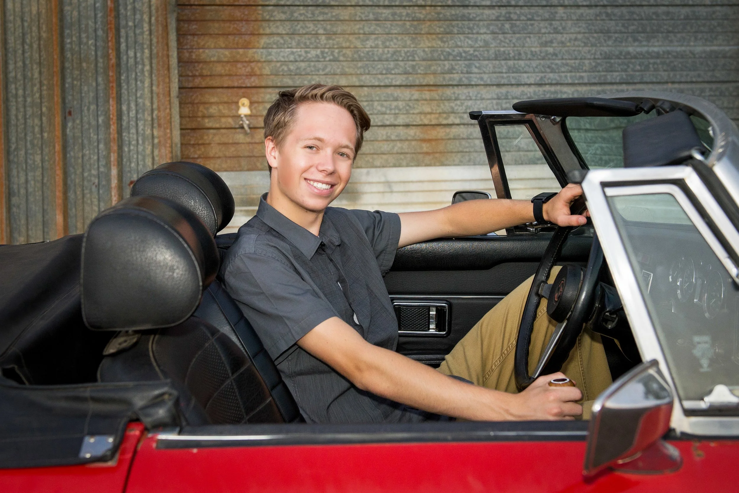 A young man with a smile sitting in a vintage red convertible car, with his left arm resting on the door and his right hand on the steering wheel, in front of a corrugated metal garage door.