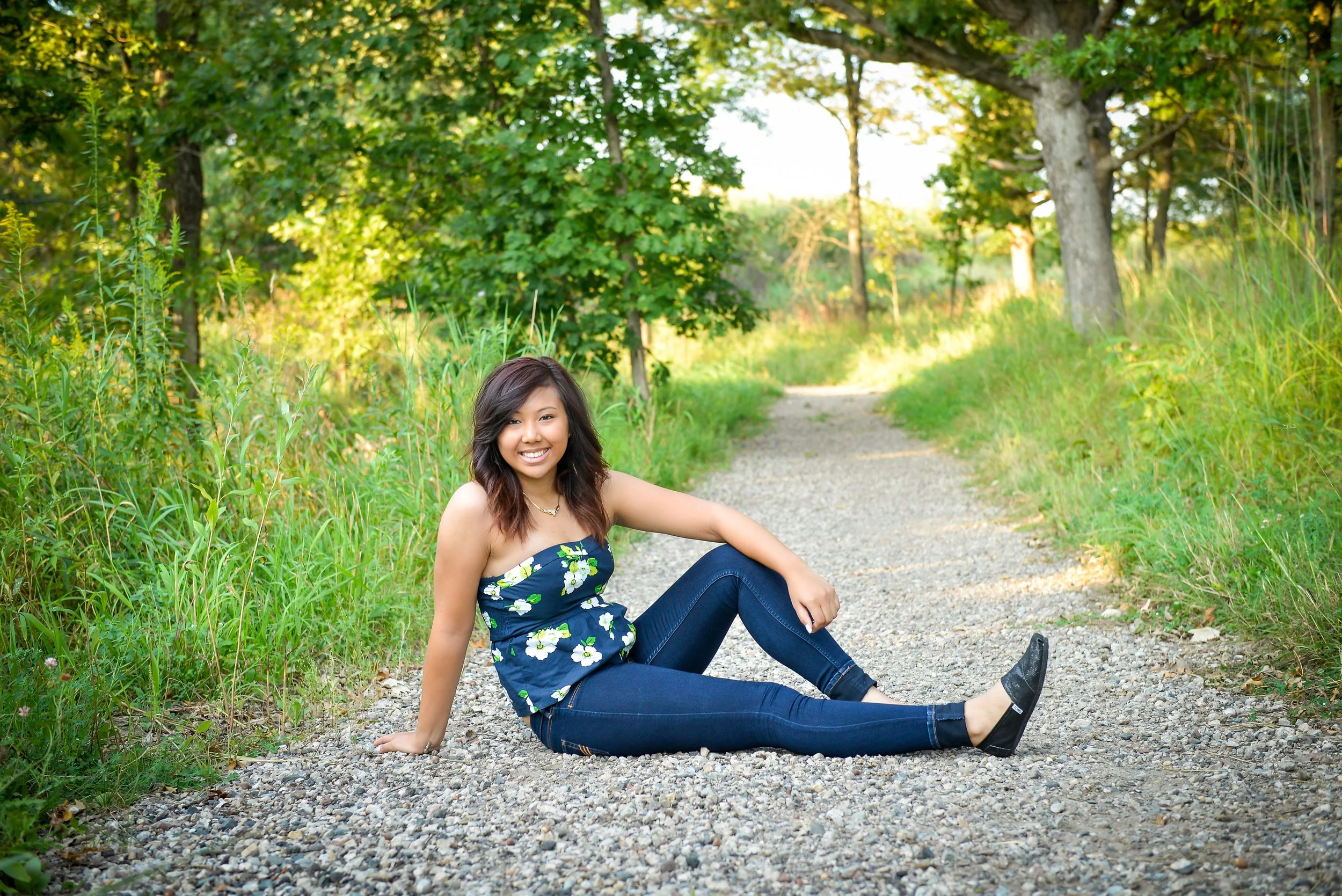 A young woman sitting on a gravel path in a lush green park, smiling at the camera, wearing a strapless floral top and jeans.