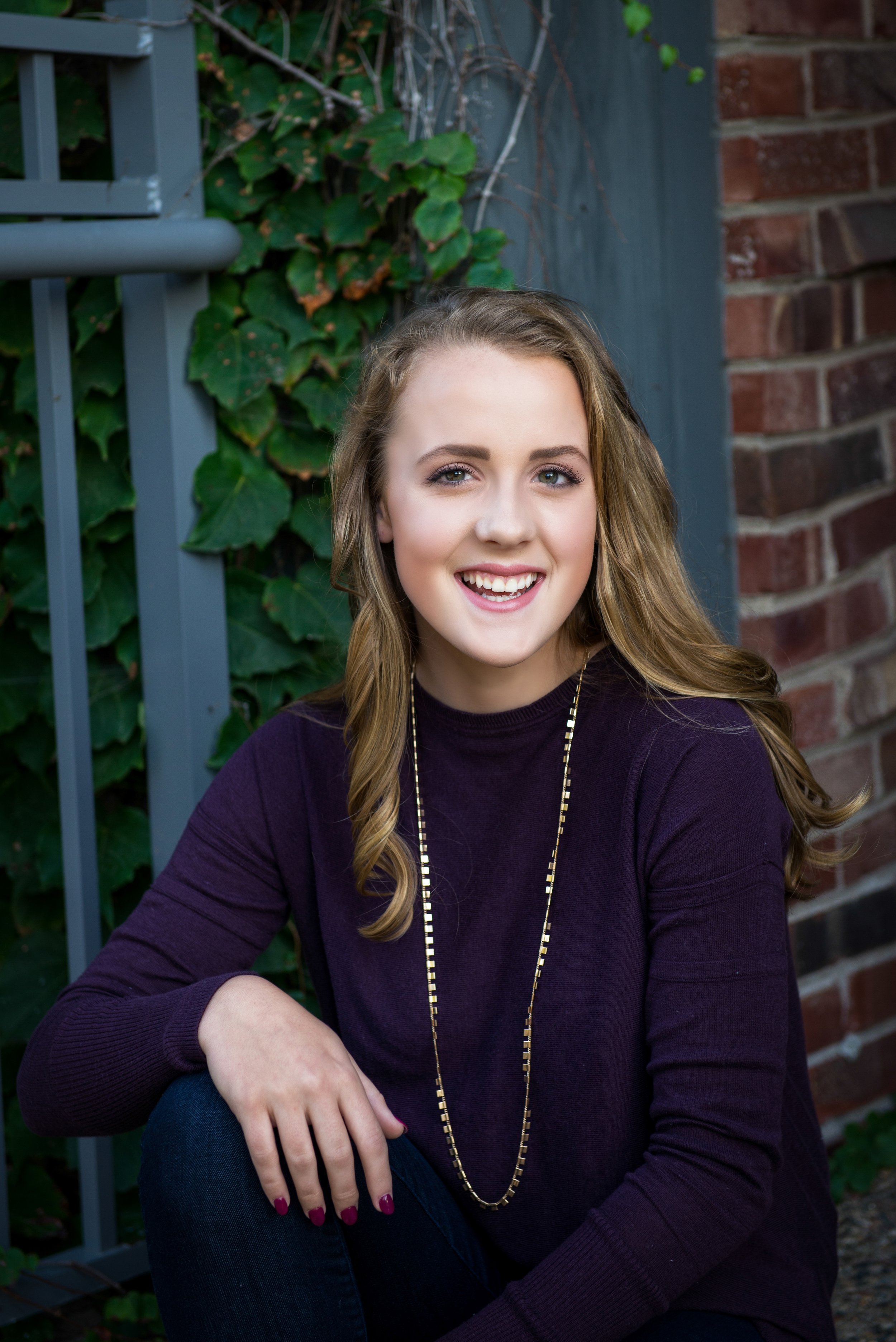 A young woman with long, wavy blonde hair smiling while sitting outdoors near a brick wall and green ivy plants.