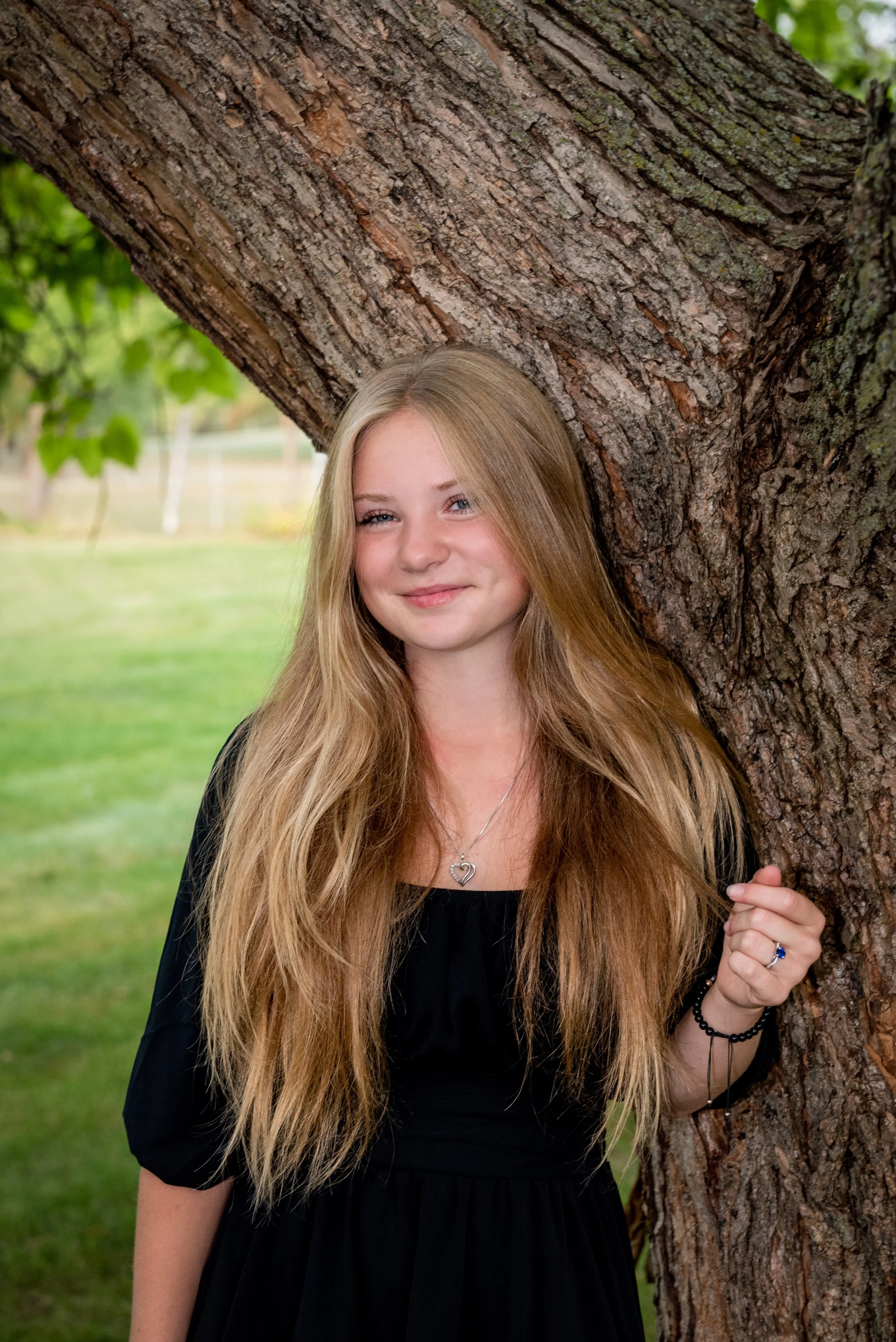 A young woman with long blonde hair smiling and leaning against a large tree outdoors.