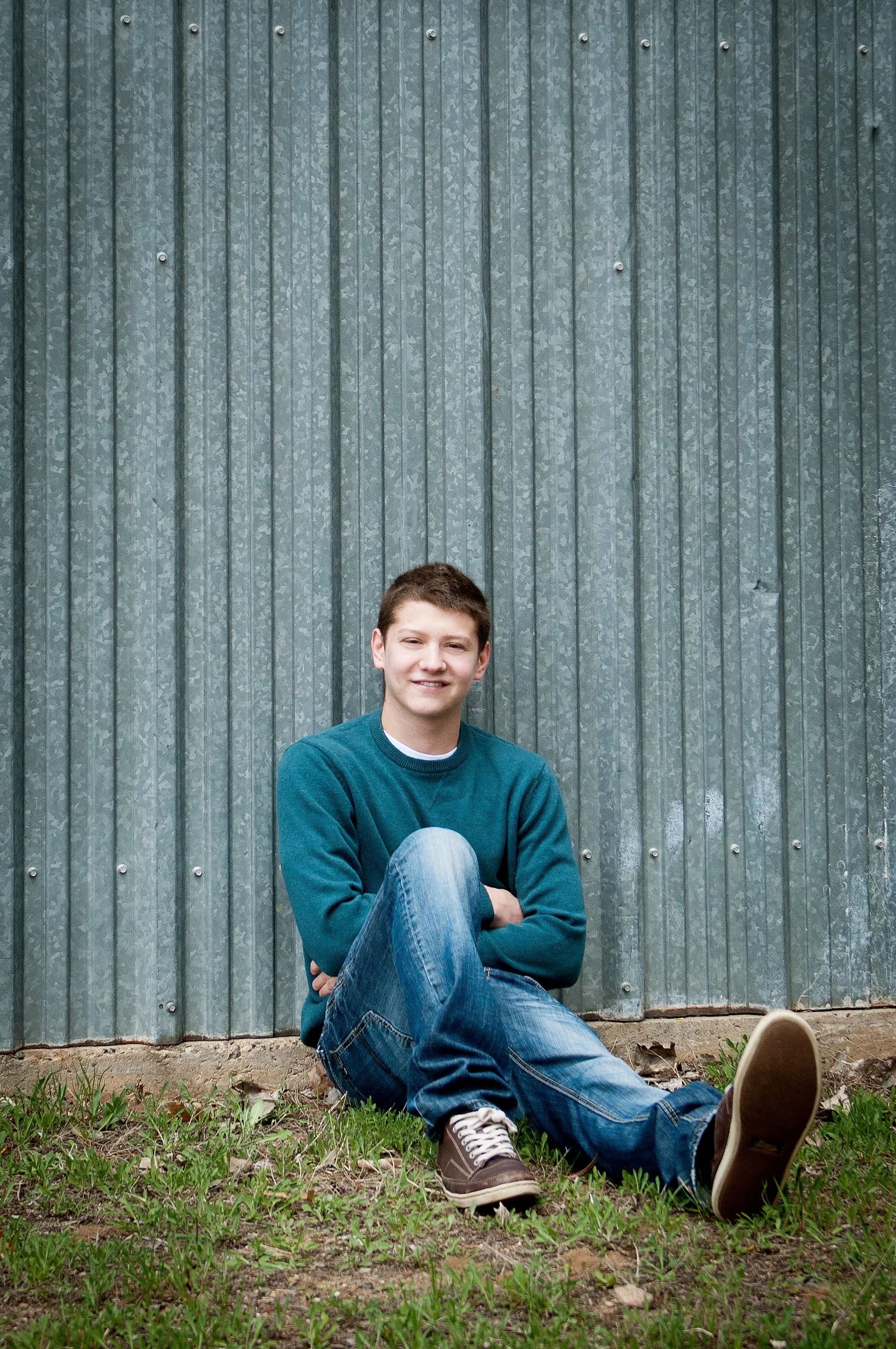 A teenage boy sitting on grass in front of a corrugated metal wall, smiling.