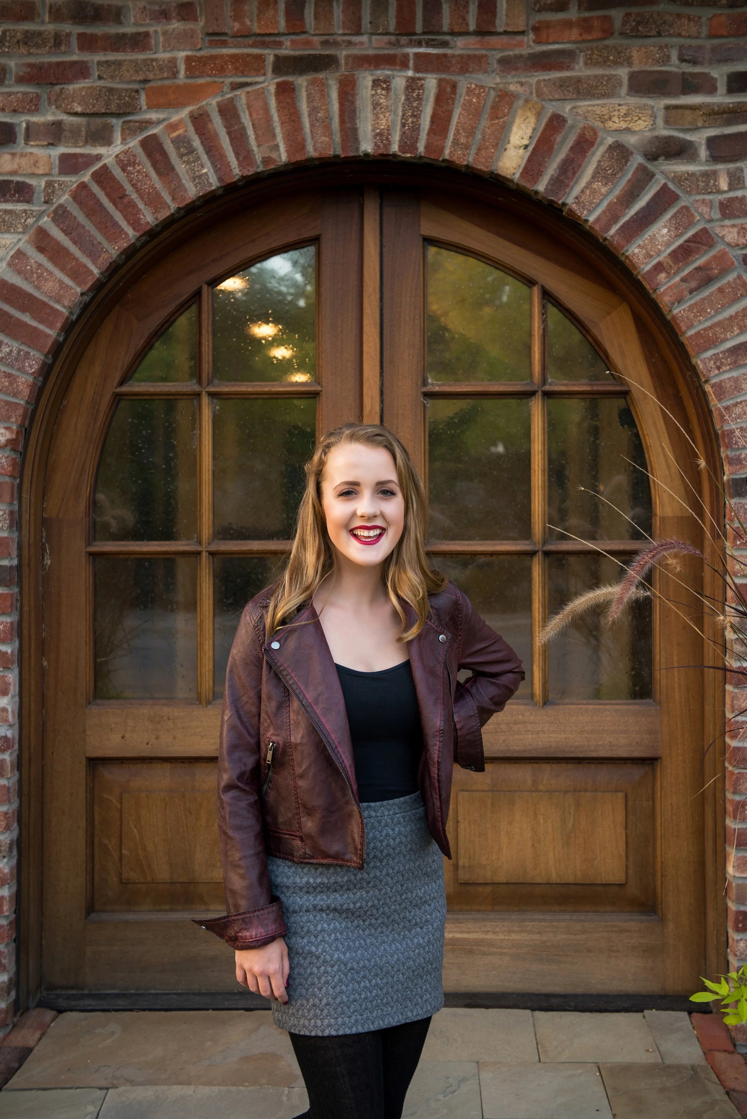 A woman with long wavy hair wearing a brown leather jacket, black top, and gray skirt, standing in front of a wooden arched window.