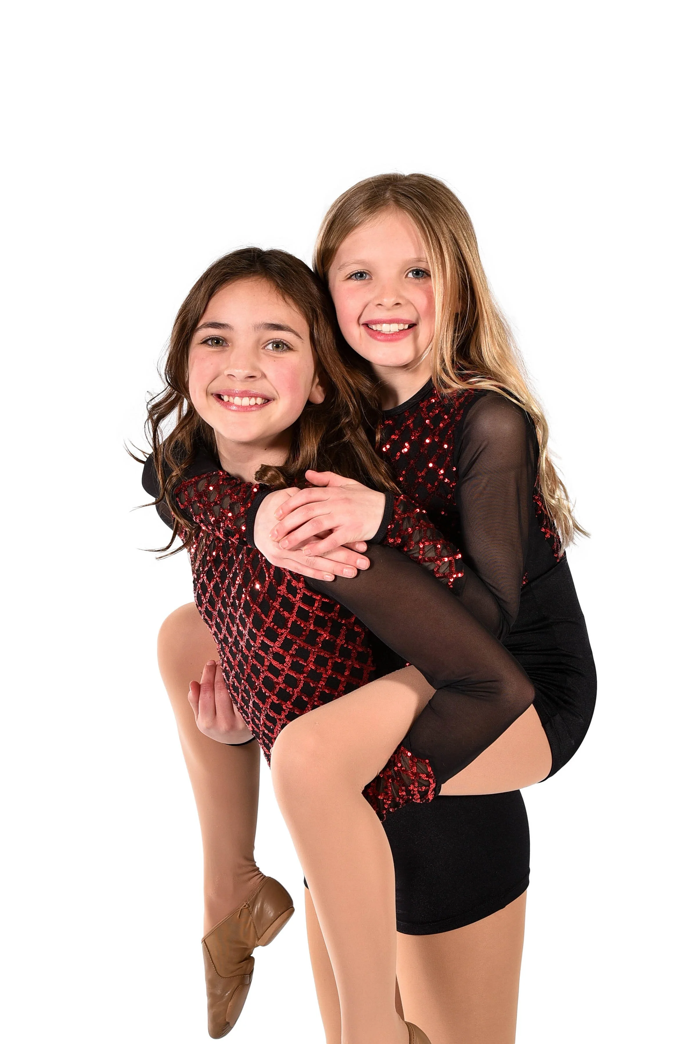 Two young girls in black and red dance costumes smiling, one giving a piggyback ride on a white background.