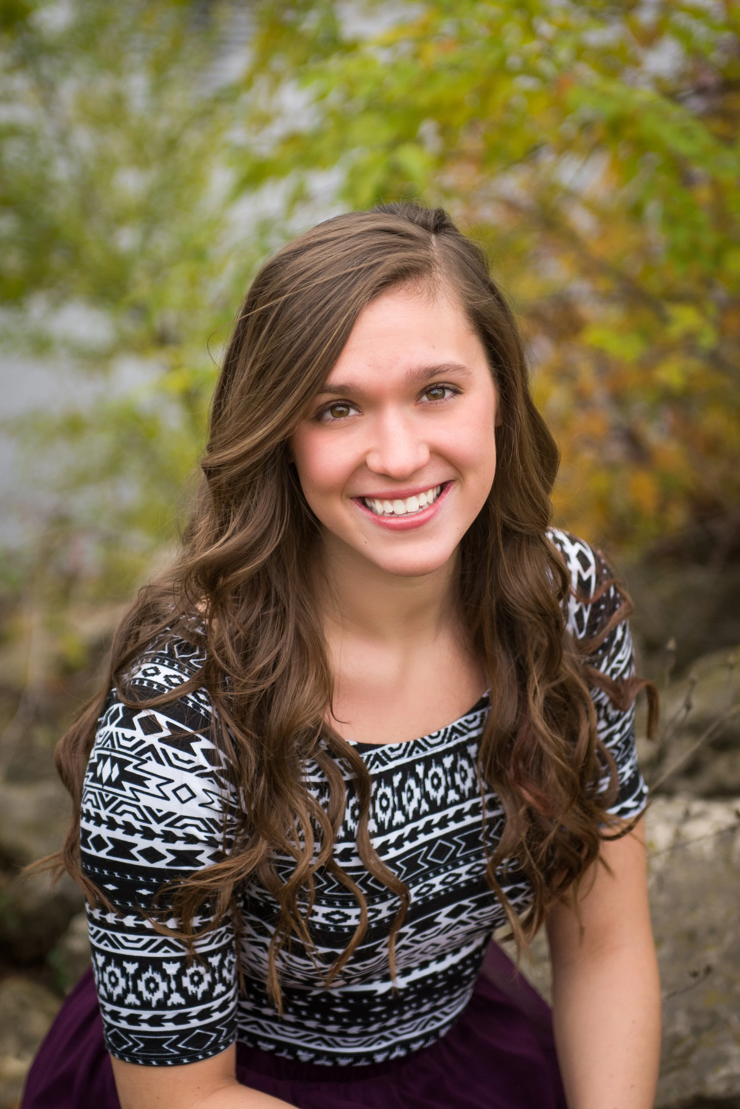 A young woman with long, wavy brown hair smiling outdoors, wearing a black and white patterned top and a dark skirt, with green and yellow autumn leaves in the background.