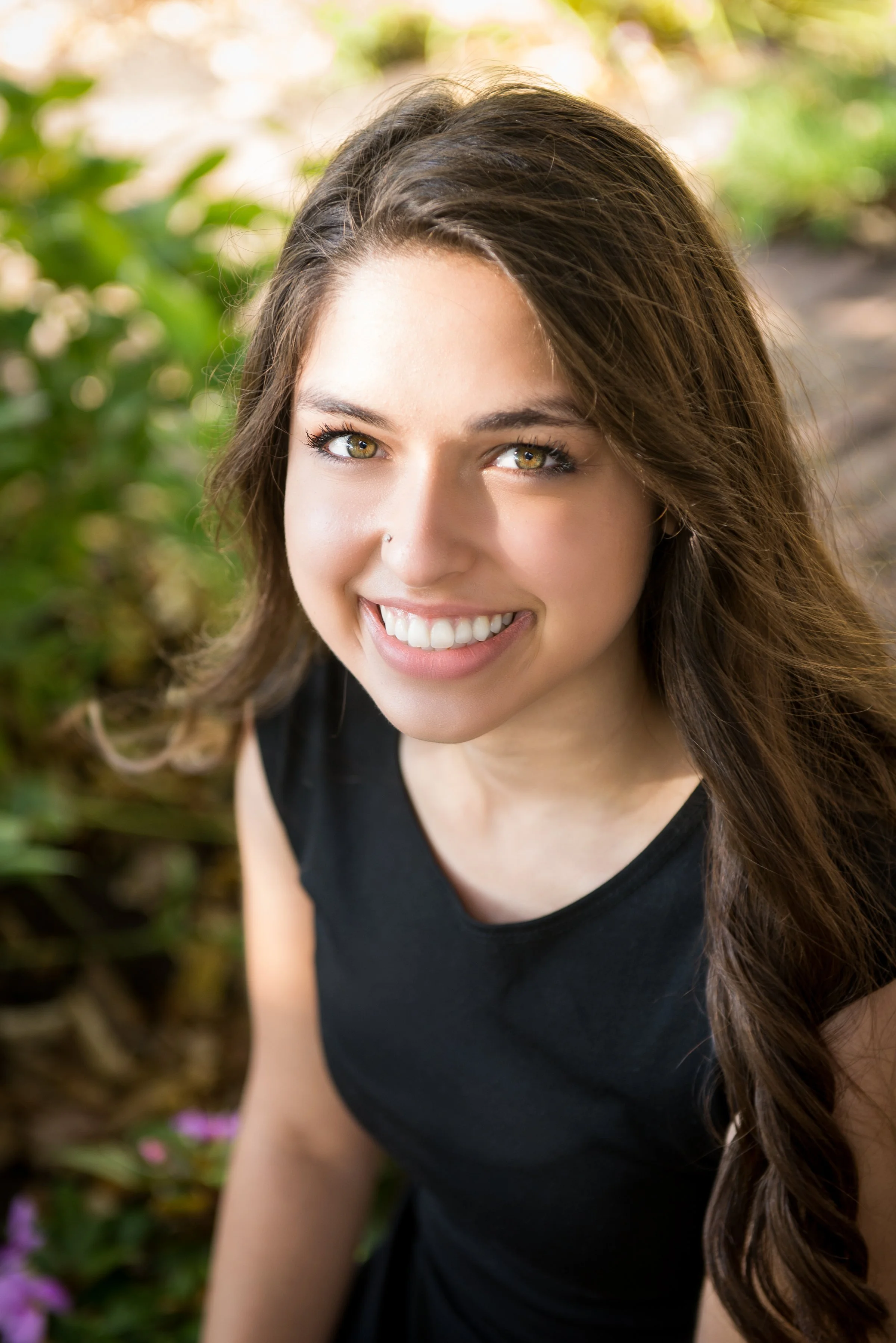 Smiling young woman with long brown hair and green eyes, wearing a black sleeveless top outdoors with greenery in the background.