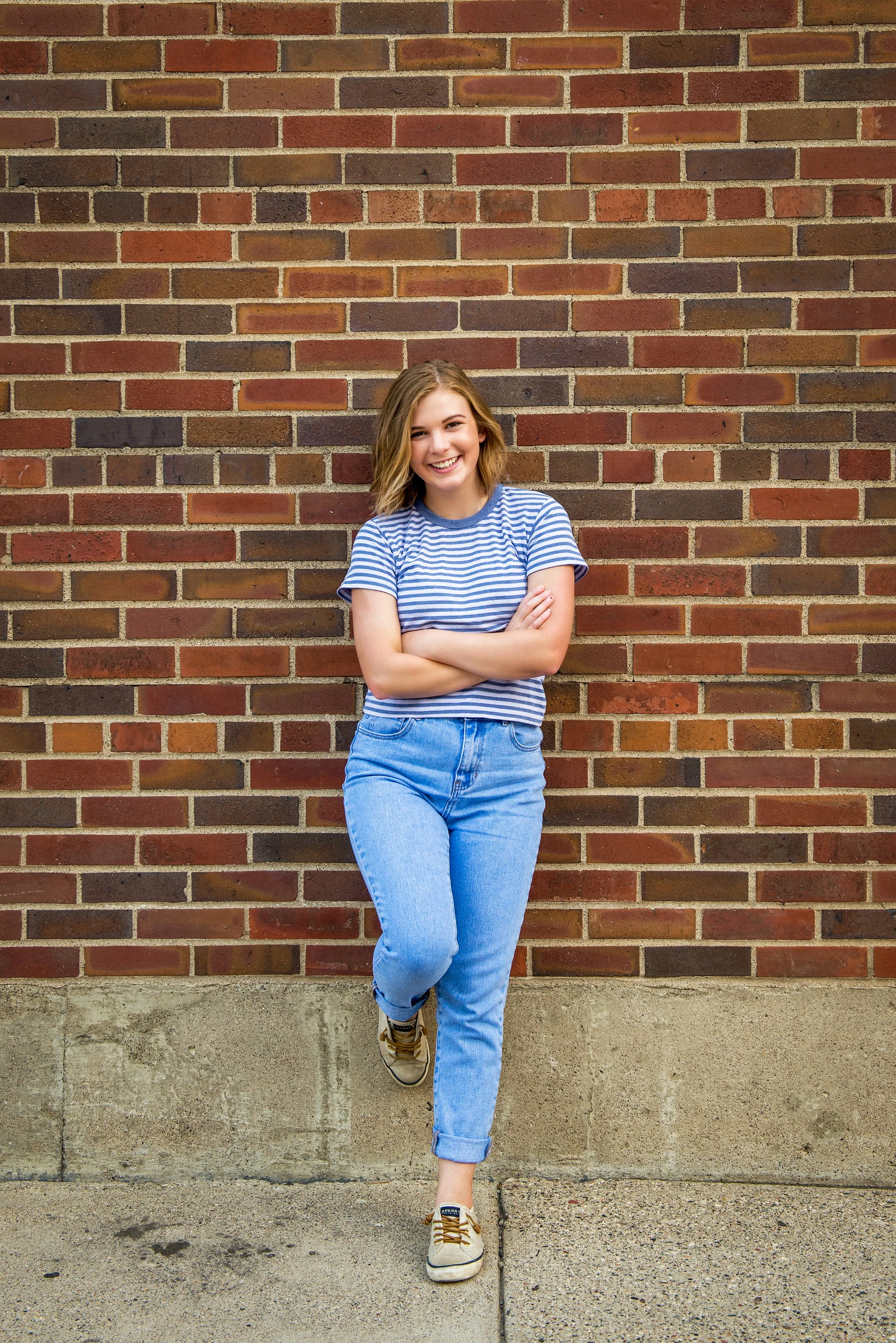 Young woman with shoulder-length wavy hair, wearing a blue and white striped T-shirt, blue jeans, and sneakers, standing against a brick wall with arms crossed, smiling.