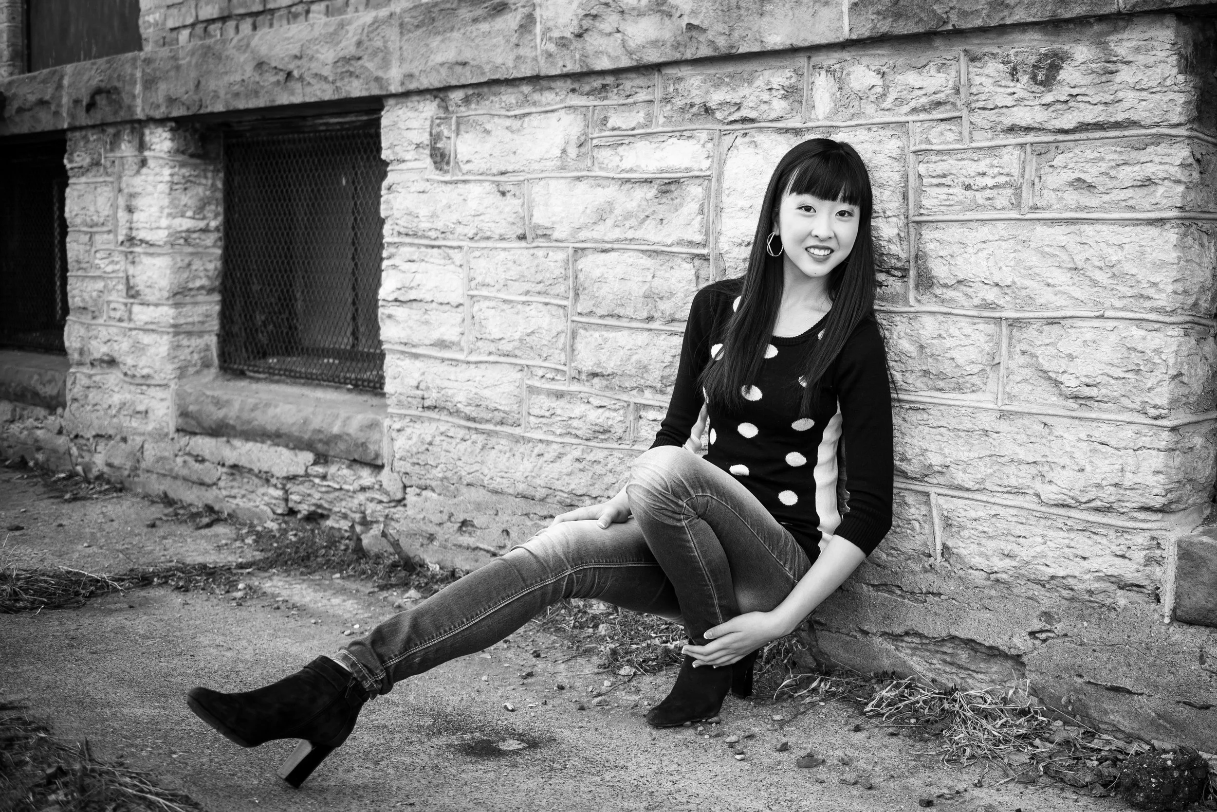 A woman with long dark hair wearing a polka dot sweater, jeans, and ankle boots sitting on the ground against a brick wall, smiling.