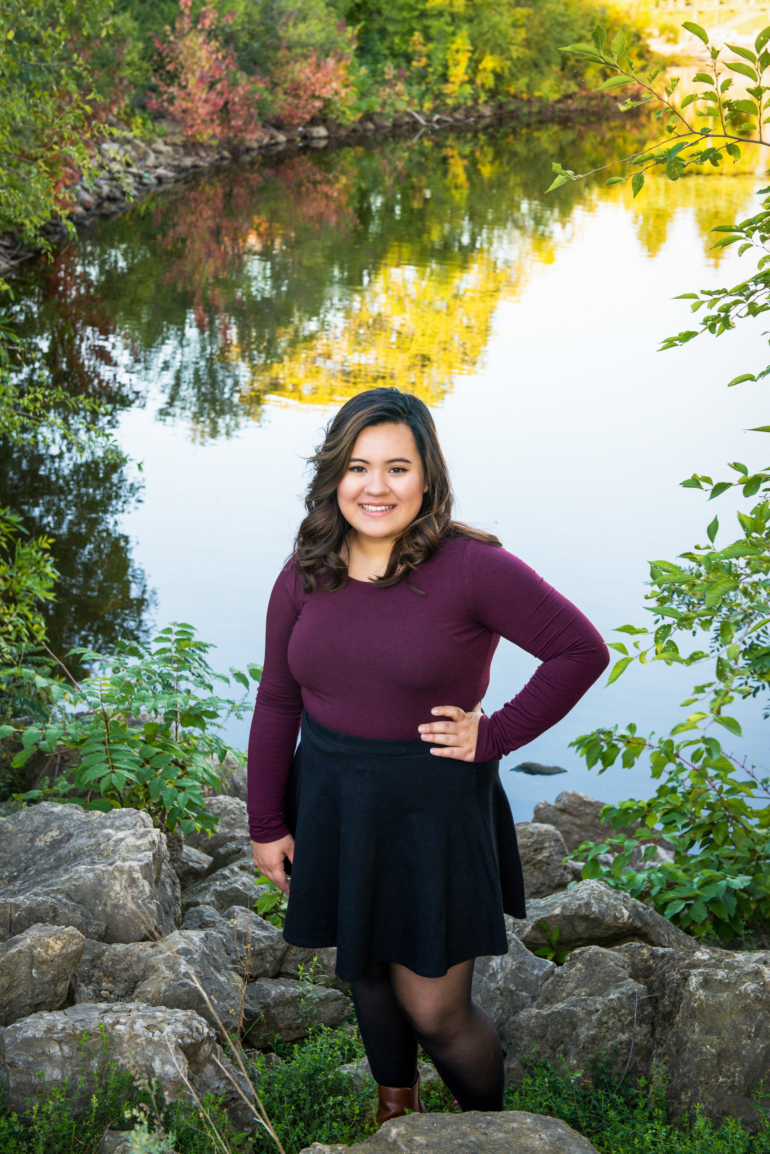 A woman smiling and standing among rocks near a river, surrounded by green trees with fall foliage, during daytime.