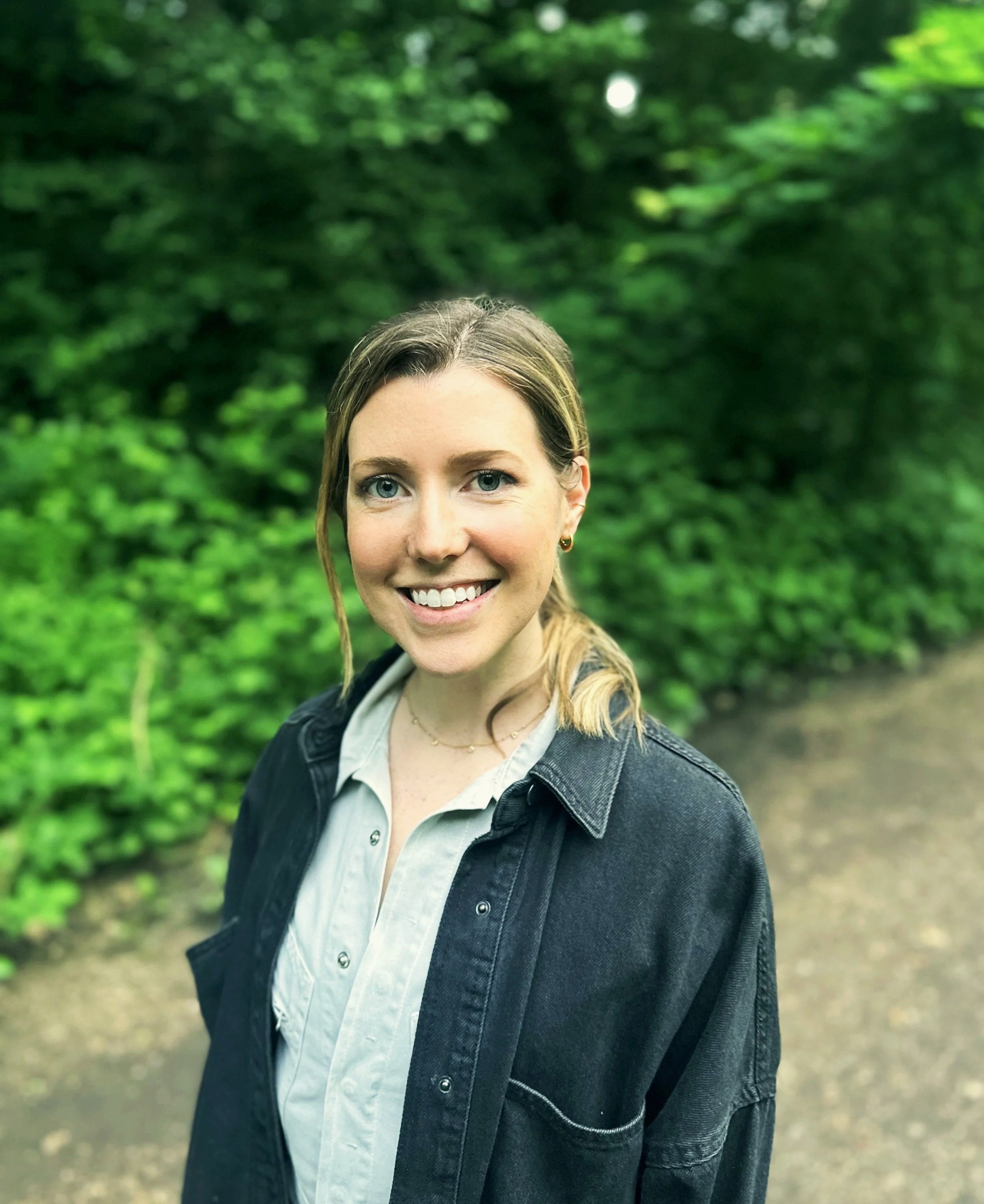 Smiling woman searing a white button down shirt under a black colored shirt, on a path in front of a green background. 