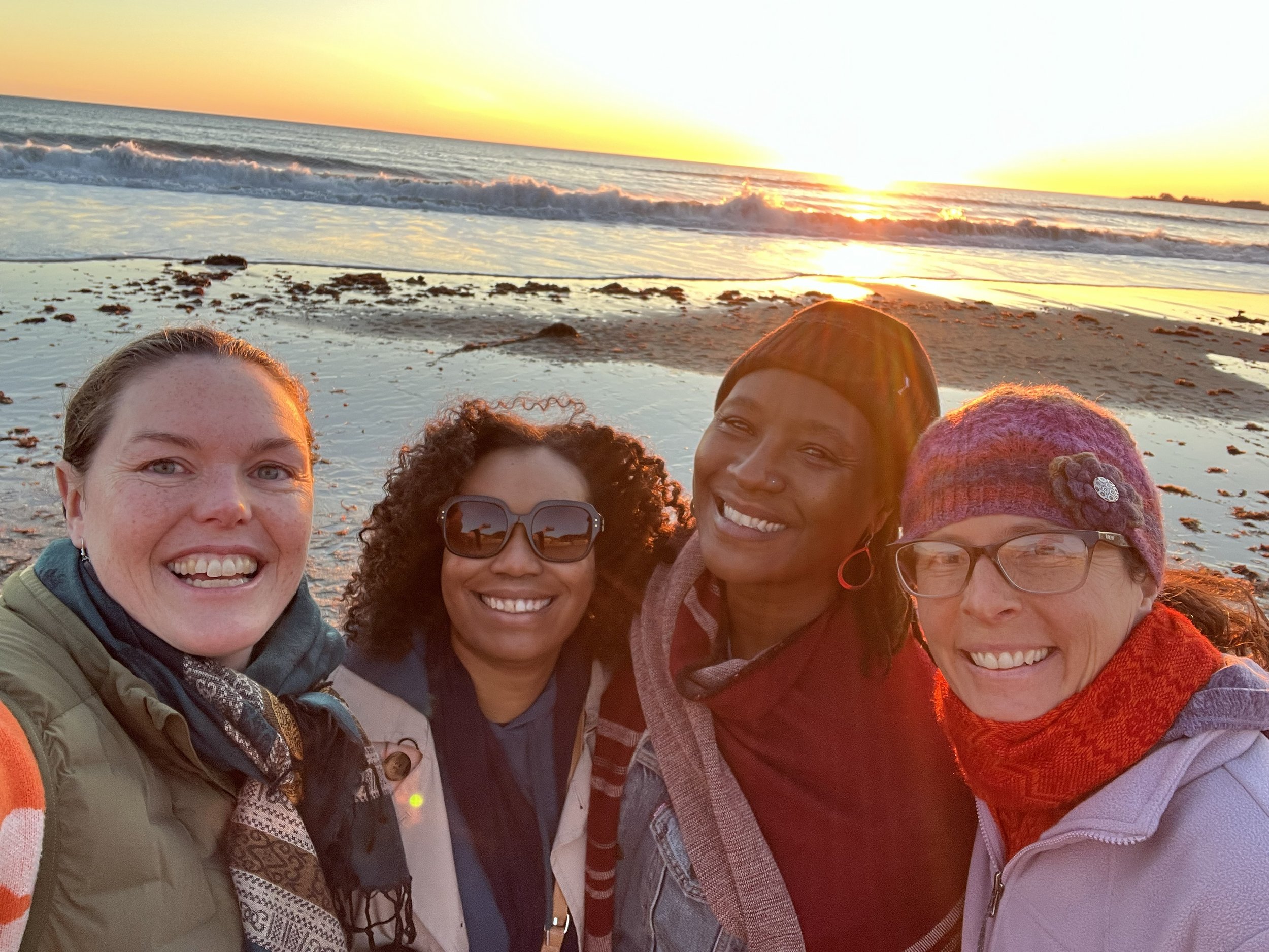 Four women smiling at the beach during sunset with the ocean and waves in the background.