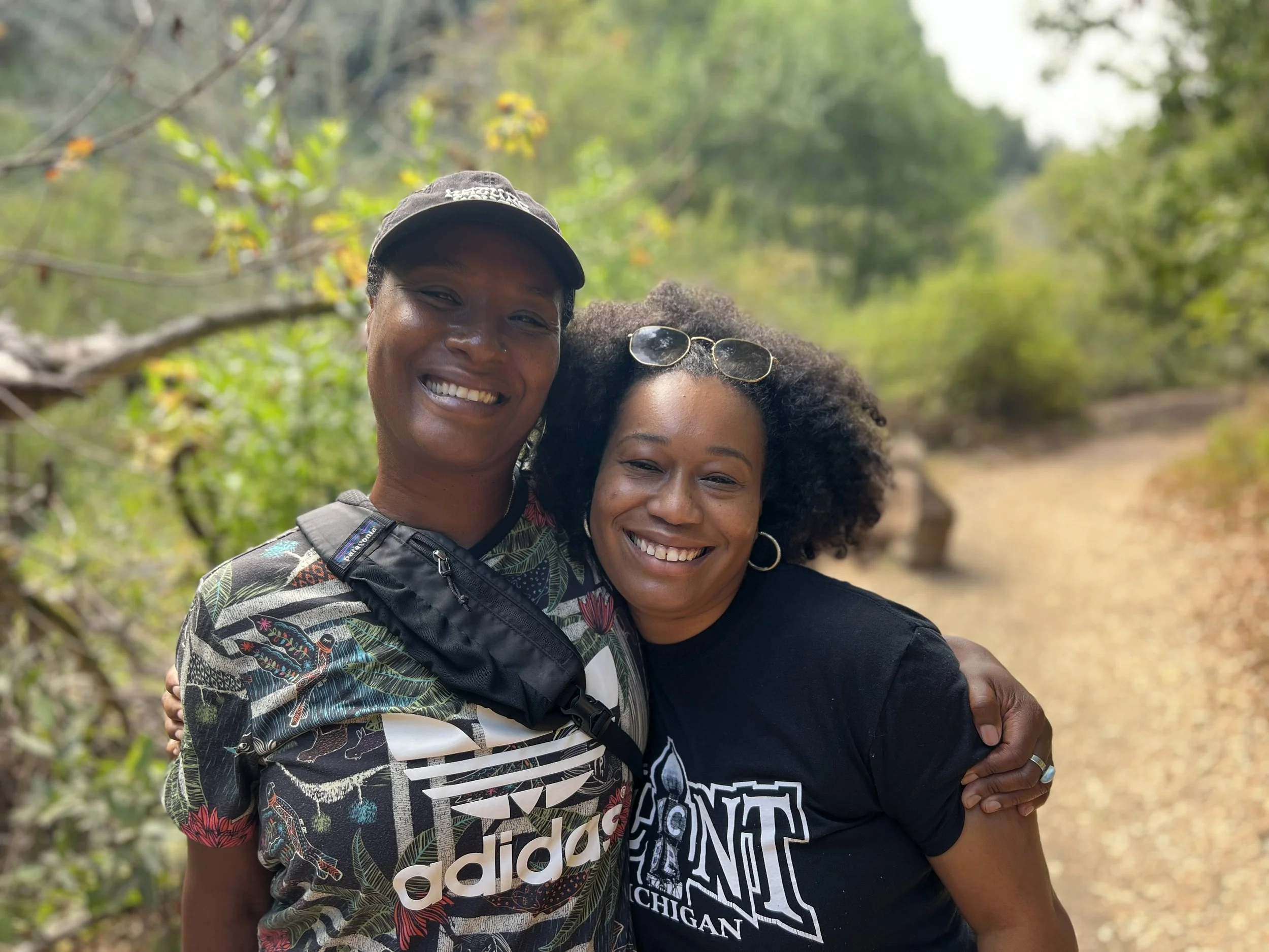 Two women smiling with their arms around each other on a nature trail surrounded by trees and greenery.