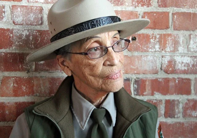 A woman wearing glasses, looking off into the distance, wearing a National Park Service uniform with a collared shirt, a jacket, a tie and a hat, standing in front of a brick wall.