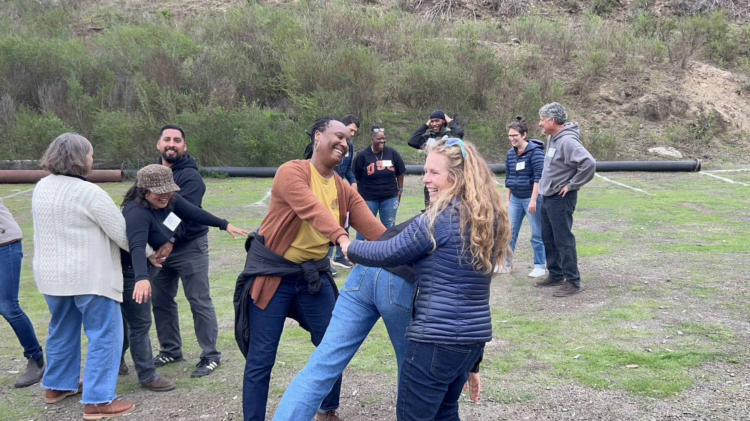 Group of adults outside, smiling, in groups of three, with two partners holding hands and one person in the middle. Several adults are laughing in the background. 
